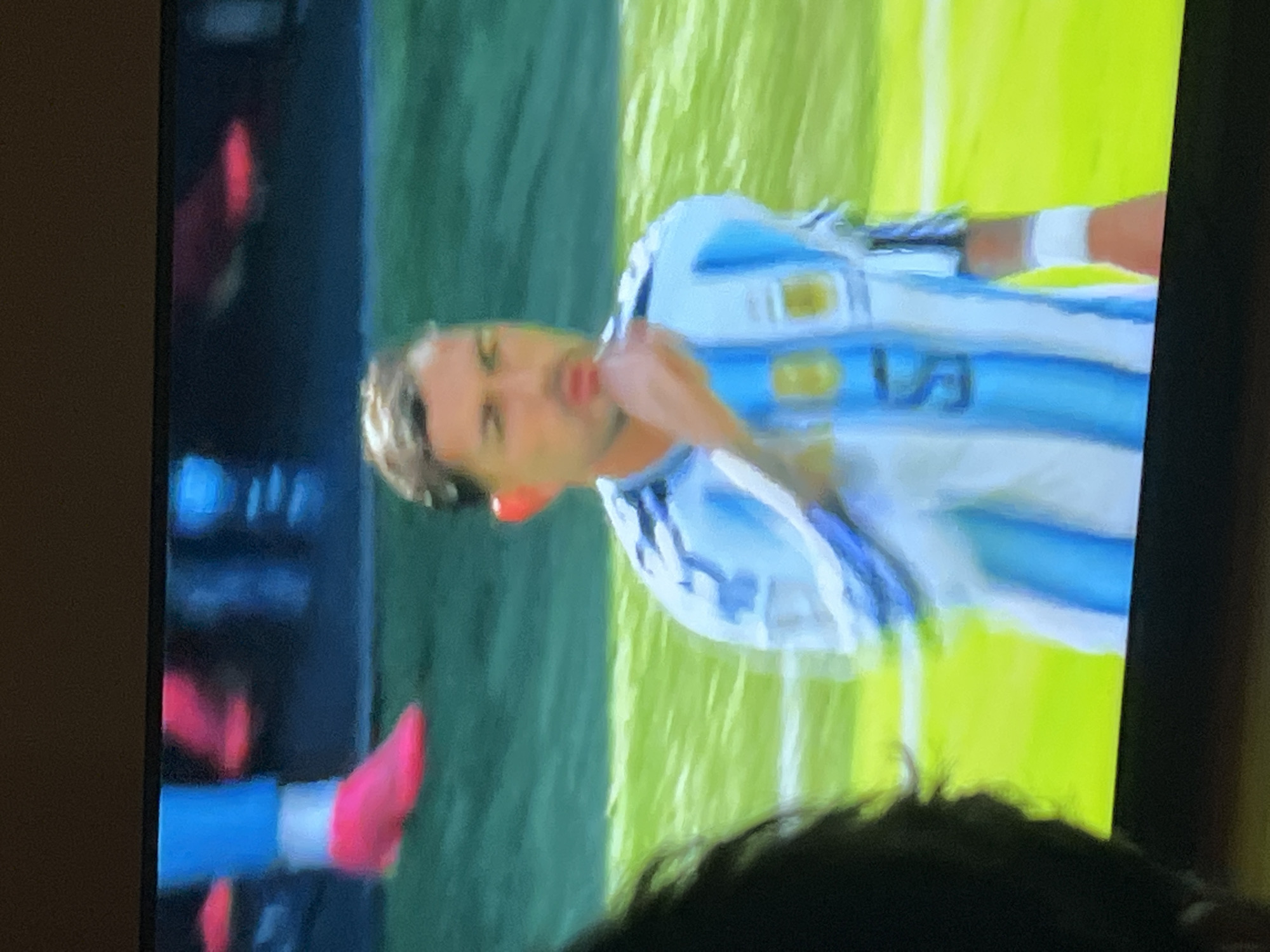 The image shows a man wearing an Argentinian soccer jersey with white and blue stripes. He has dark hair and is looking to the side with his hand near his mouth. The jersey has a gold emblem and the number "5" is visible on the front. He is also wearing a white wristband. The background appears to be a soccer field with green grass and a dark backdrop, possibly a stadium. The image is blurry and appears to be taken from a screen. There are also other blurred figures and elements visible in the background, including what appears to be a person's legs in bright pink shoes and blue socks in the upper left corner. The overall scene suggests a sporting event. There is no information in the image to suggest it is located in Moscow, Russia.