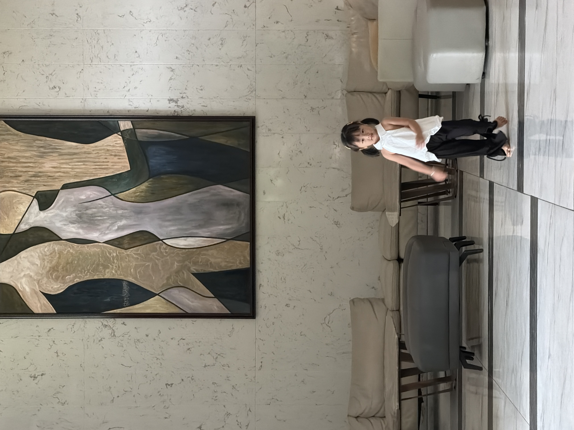 The image depicts a young girl with dark hair tied in pigtails, wearing a white halter top and black pants, standing on a marble floor. She is in the middle of a room with light-colored marble walls. In the background, there is a large abstract painting featuring stylized figures in muted colors, framed in dark wood. The room is furnished with modern seating, including a beige sectional sofa and a dark gray ottoman with multiple legs. The lighting is bright, suggesting an indoor setting, possibly a lobby or a waiting area. The girl appears to be in motion, with her right arm extended and her body tilted as if she is dancing or turning. The overall aesthetic is contemporary and minimalist. Without further information, the specific location within Nha Trang, Vietnam, cannot be determined from the visual evidence alone.