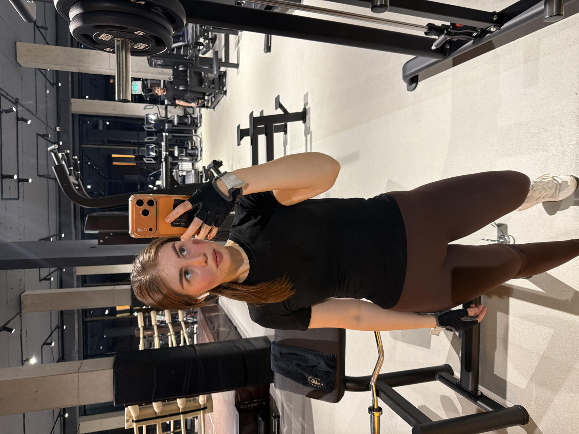A young woman is taking a selfie in a gym, located in Усады, Russia. She is wearing a black t-shirt, brown leggings, and gym gloves. She is holding a smartphone with an orange case. The gym equipment visible includes weight machines and a pull-up bar. The floor is a light gray color.