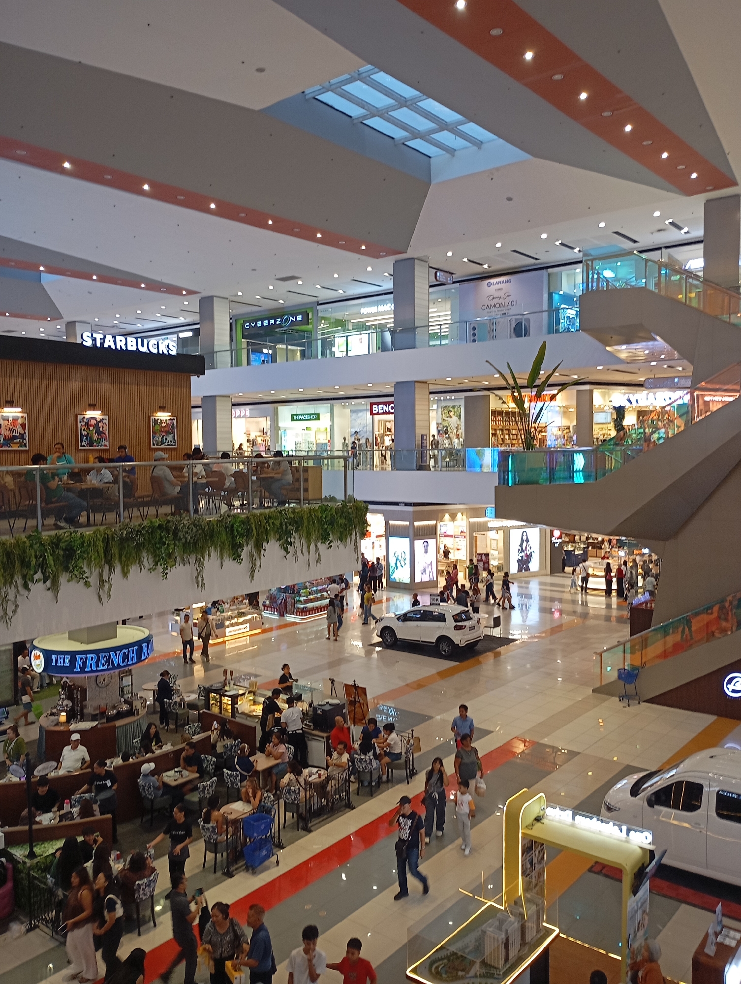 This image captures a lively, multi-level indoor shopping mall in Davao City, Philippines, viewed from an elevated perspective. The mall boasts modern architecture with high ceilings, overhead lighting, and skylights that provide natural illumination, complementing the artificial lights.
Numerous people are seen across different levels, engaging in various mall activities. Many are walking, while others are dining and socializing in cafes and restaurants. On an upper level, a prominent Starbucks cafe, identifiable by its sign and distinct wooden facade, is bustling with patrons seated at tables, some of whom are visible through a railing adorned with cascading greenery.
On the ground floor, another busy dining area featuring "The French Baker" with a circular counter is visible, surrounded by customers. Further down, several people are gathered around various tables. A white SUV is prominently displayed on the ground level, potentially as a promotional exhibit, and a smaller architectural model display with glowing yellow accents is also present.
Retail stores line the mall's interiors, with visible signage for "CYBERZONE" and "BENC" (likely Bench). A large banner on an upper floor advertises "CAMON 201" and "LAHANANG," possibly for a mobile phone. Other partial texts like "SHOPEE" and "OPENING SOON" are also discernible. The atmosphere appears casual and energetic, consistent with a busy daytime shopping and leisure destination.