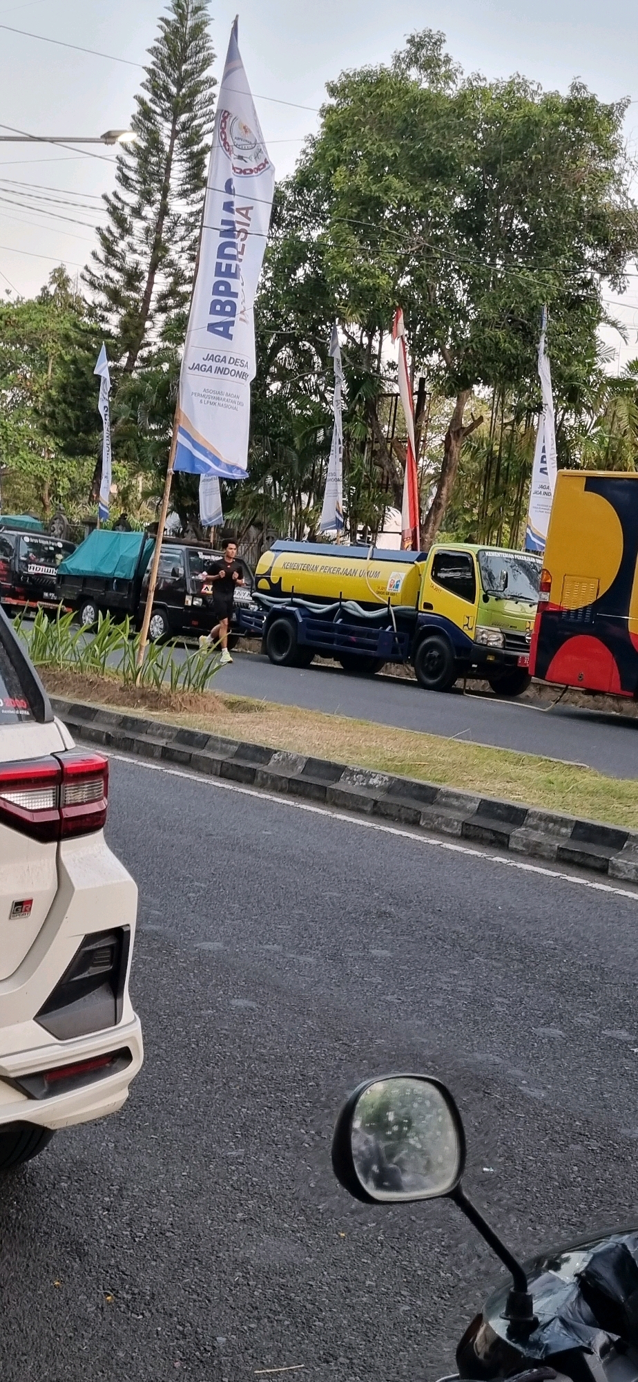 A man runs along a street in Denpasar, Indonesia, with flags and vehicles in the background. The man is wearing a black t-shirt and black shorts. To his left, a yellow water truck with the text "KEMENTERIAN PEKERJAAN UMUM" is parked. Behind the truck, a yellow and red painted vehicle is visible. In front of the man, several black pick-up trucks are parked. Tall trees and flags bearing Indonesian text line the side of the road. In the foreground, the rear of a white car and a motorcycle's side mirror are visible. The scene appears to be a street event or parade.