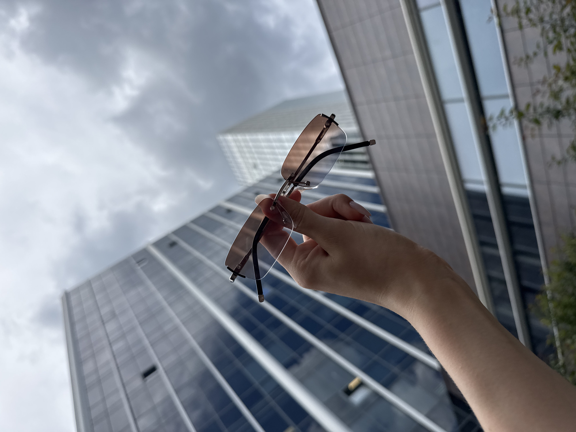 A person's hand holds a pair of rimless sunglasses with gradient brown lenses in front of a blurred background of modern glass-fronted buildings and a cloudy sky, likely in a city like Yekaterinburg, Russia. The buildings appear to be high-rises, with reflective surfaces and horizontal banding. The focus is on the sunglasses and the hand, with the architecture serving as a backdrop.