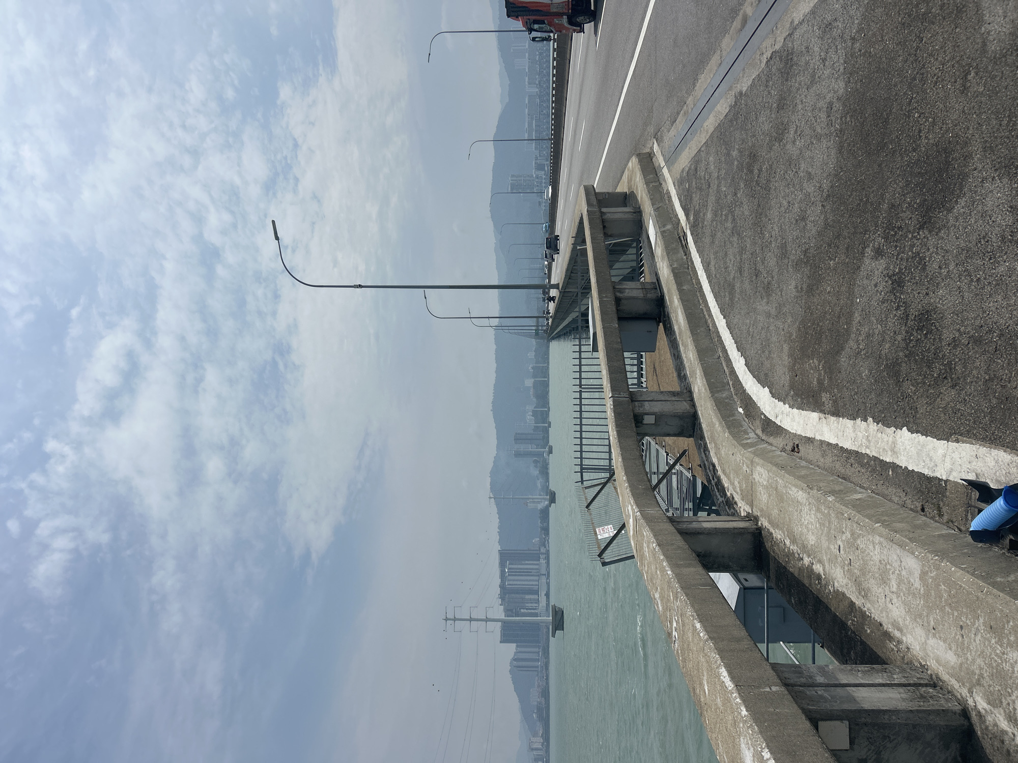 A wide-angle shot captures a view from a concrete bridge on a cloudy day. The bridge features a concrete barrier along the left side, overlooking a body of water with waves. Beyond the water, a city skyline is visible with buildings and power lines. The bridge's roadway, with multiple lanes and a red truck, curves to the right in the distance. The sky is filled with scattered white clouds.