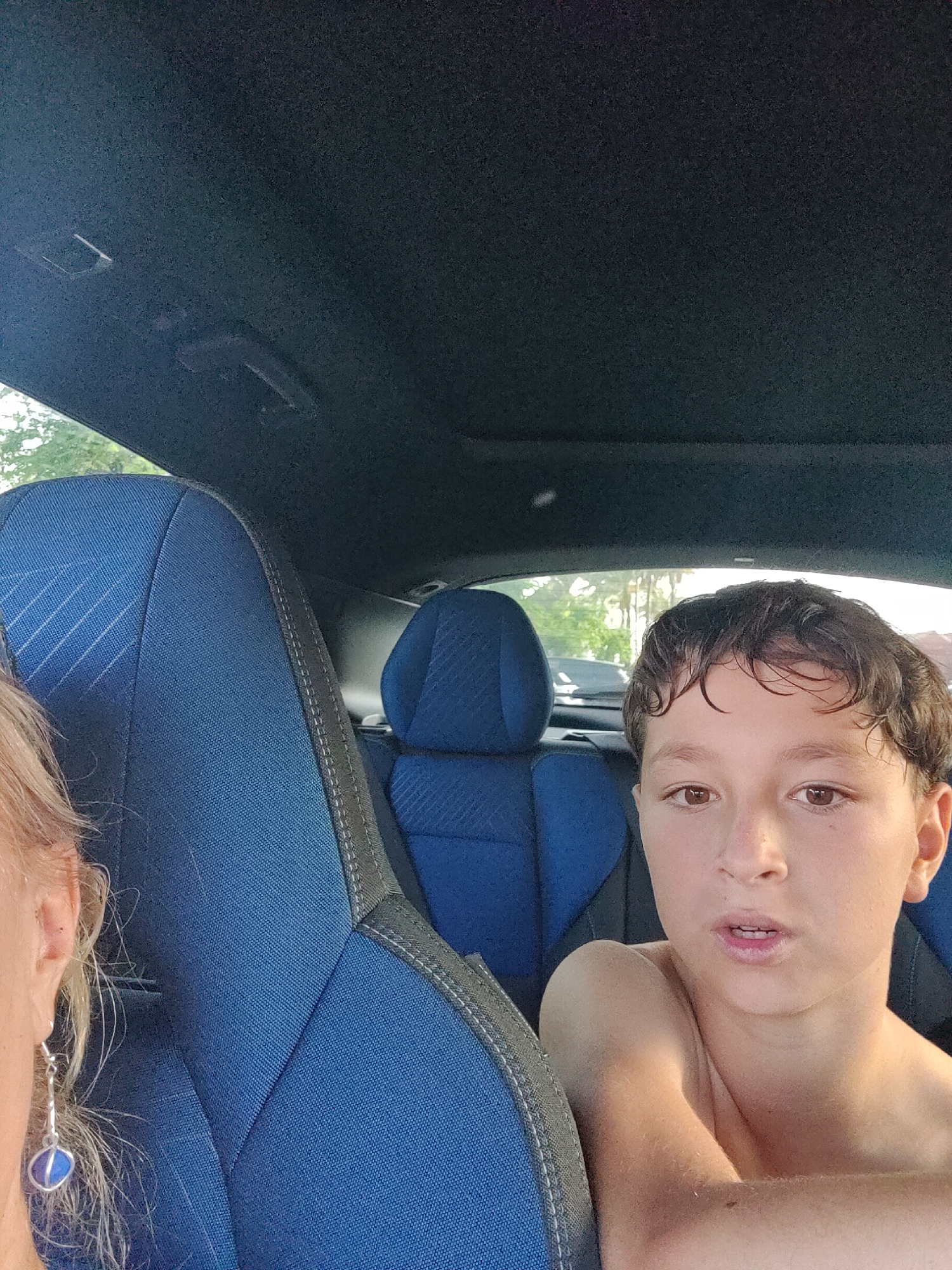 A boy is in the passenger seat of a car, looking directly at the camera with a neutral expression. He has brown hair and brown eyes. His bare shoulders are visible. A woman's shoulder and ear with an earring are visible on the left side of the frame. The interior of the car is dark blue with grey stitching. The sun is shining through the windows, indicating it is daytime. The background outside the car is blurred, showing trees and other vehicles.