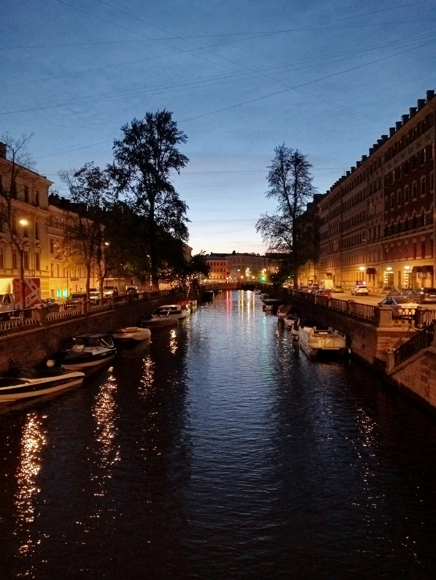 The image depicts a canal in Saint Petersburg, Russia, at dusk. The sky is a deep blue, transitioning to a lighter hue near the horizon. Several buildings line both sides of the canal, illuminated by warm, yellow streetlights. Trees with dark silhouettes are interspersed along the waterfront. Numerous boats are docked along the canal banks, their reflections shimmering on the dark water. The water itself appears calm, with ripples reflecting the lights from the buildings and the sky. The overall scene conveys a peaceful and atmospheric urban evening.