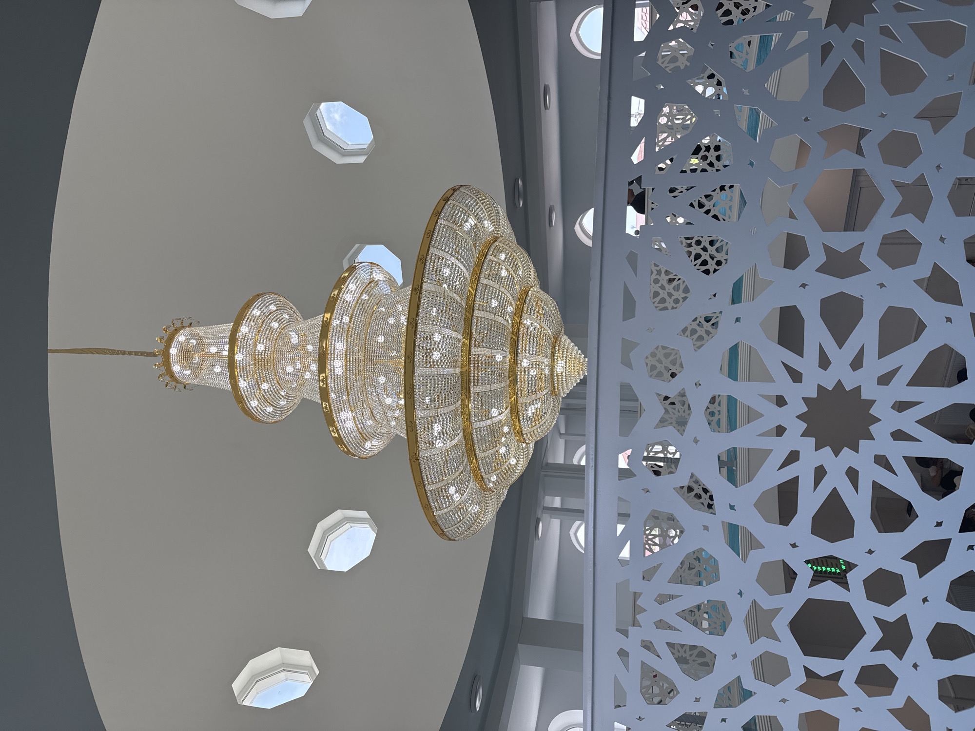An ornate, multi-tiered crystal chandelier with gold accents hangs from a domed ceiling in what appears to be a mosque or large public building in Novaya Adygeya, Russia. The ceiling has several octagonal skylights showing a blue sky with some clouds. Below the chandelier, a white, intricately patterned screen with geometric Islamic motifs is in the foreground. Through the screen, glimpses of the interior can be seen, including arched windows, white walls, and the tops of people's heads. The overall impression is one of grandeur and a blend of traditional and modern architectural elements.