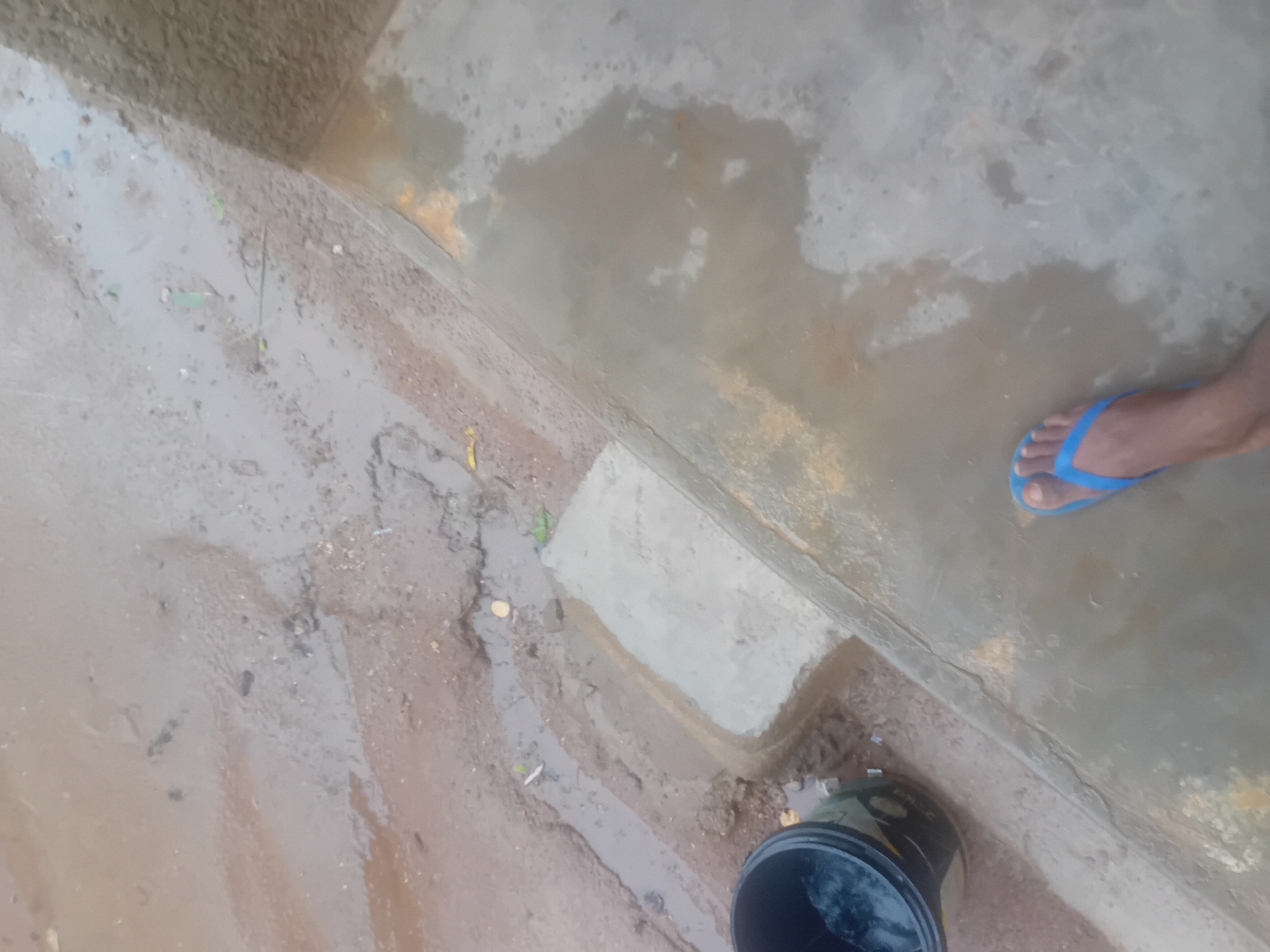In Jauro Yinu, Nigeria, a close-up shot reveals a flooded outdoor environment. The ground is a mixture of mud and water, with visible puddles and a small stream flowing. A black bucket, partially filled with water, sits to the left of the frame. In the foreground, a person's foot, wearing a bright blue flip-flop, is visible on a wet, concrete-like surface. The scene depicts an area affected by recent rainfall or water accumulation.