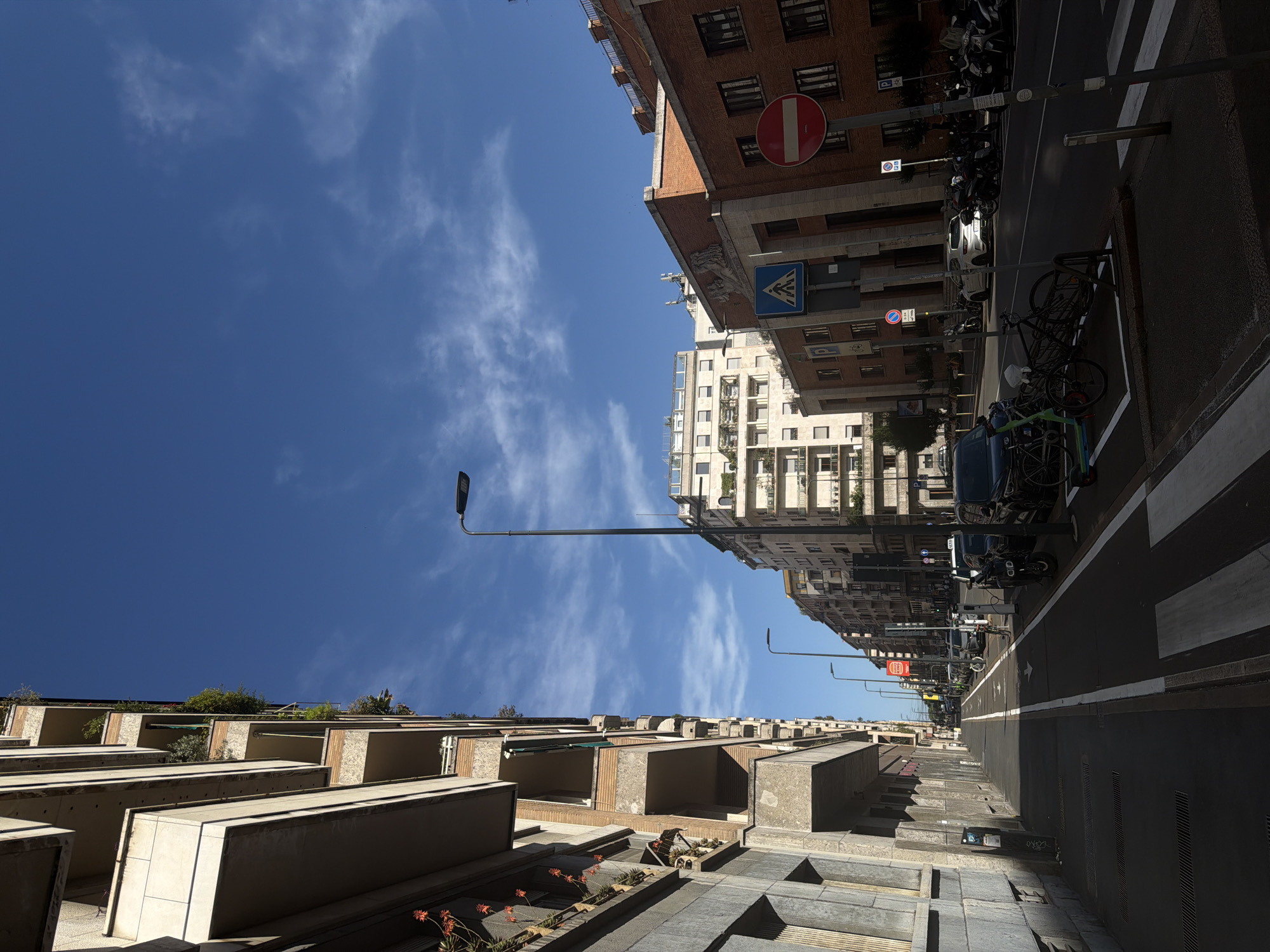 The image depicts a street scene in Milan, Italy, on a clear day with a bright blue sky and scattered white clouds. The perspective is from a sidewalk, looking down a street lined with apartment buildings and other structures.
On the left side of the street, a tall apartment building with multiple balconies is prominently featured. The balconies have various plants and some flower arrangements, indicating residential occupancy. The building has a textured facade, appearing to be a mix of stone and painted surfaces.
Across the street, several buildings of varying architectural styles and heights are visible. Some are modern with white facades and large windows, while others are older brick buildings with more traditional designs. One building has a prominent bas-relief sculpture above its entrance.
The street itself has designated lanes for vehicles and a separate lane or path marked for bicycles and scooters, as indicated by white lines. Several parked cars, motorcycles, and scooters are visible along the roadside. A few bicycles are also present, including one in a bike rack and an electric scooter.
There are traffic signs, including a red circular "no entry" sign, a pedestrian crossing sign (a blue square with a white pedestrian symbol), and parking signs. A street lamp stands near the center of the road. The sidewalk has a zebra crossing, indicating a pedestrian passage.
No people are clearly visible in the immediate foreground. The overall impression is of an urban environment, likely a residential or mixed-use area in Milan, during daylight hours. The sunlight casts shadows, indicating the time of day is likely morning or afternoon.