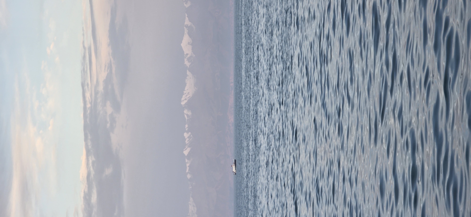 An expansive body of water, likely Issyk-Kul Lake near the city of Choktal, Kyrgyzstan, dominates the foreground with its rippling blue surface reflecting the overcast sky. A lone white boat with a dark roof is positioned in the middle distance, appearing small against the vastness of the lake. Beyond the water, a range of majestic, snow-capped mountains forms a hazy, purple silhouette against the pale sky. The clouds are layered and diffuse, with hints of pink and pale orange suggesting the soft light of either dawn or dusk. The overall impression is one of serene, natural beauty and tranquility.