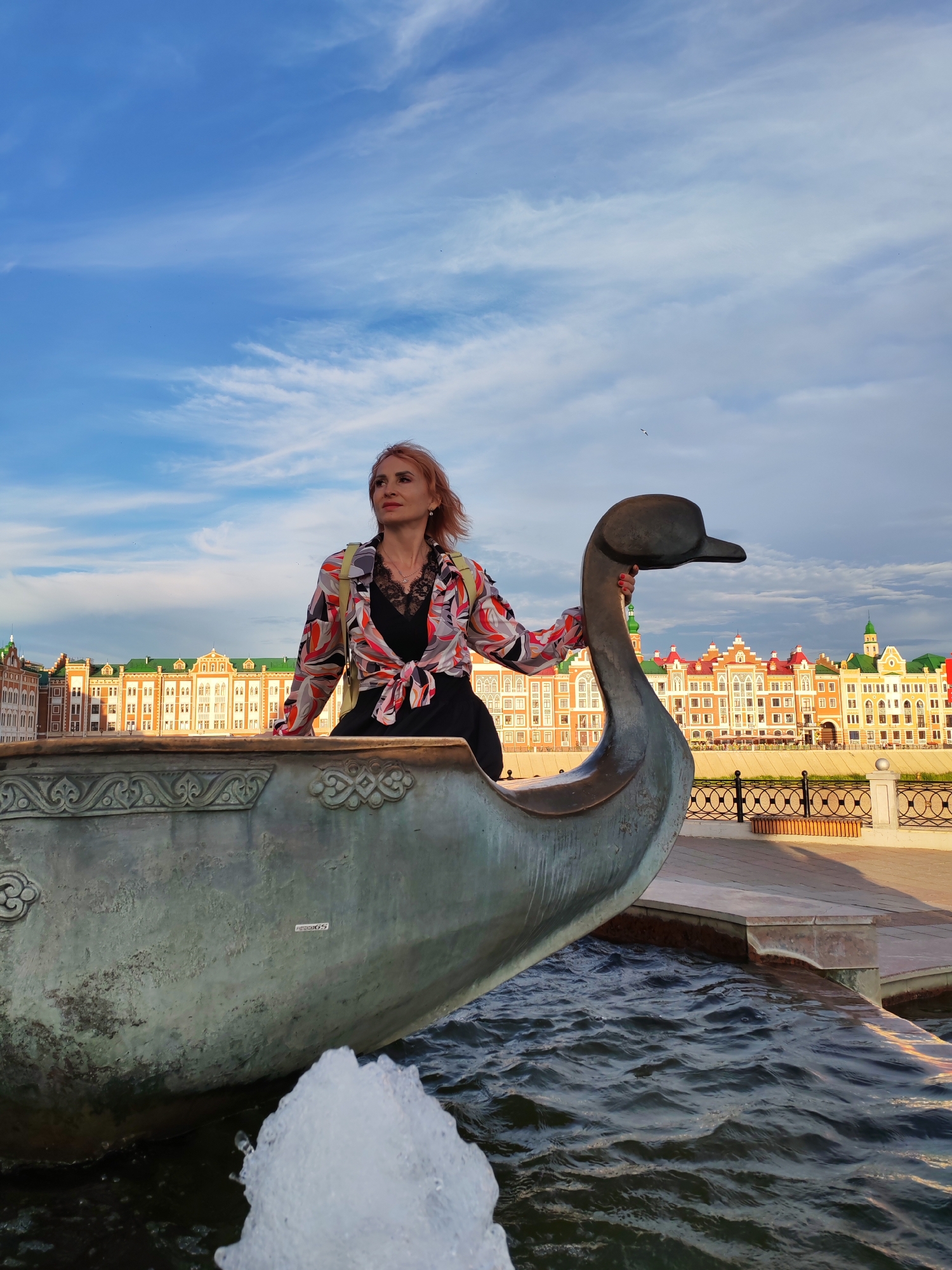 A woman with reddish-blonde hair stands in a decorative boat sculpture, which resembles a swan, with a fountain in the foreground. She is looking off to the left, with a calm and contemplative expression. The woman is wearing a black dress with a lace neckline and a vibrant, patterned button-up shirt tied at the waist. She has a light-colored backpack with yellow straps.
The boat sculpture is made of a dark, weathered metal and is partially submerged in dark, rippling water. Decorative carvings are visible on the edge of the boat.
In the background, a row of colorful, multi-story buildings with green roofs lines a waterfront promenade. A black metal fence with decorative elements separates the promenade from the buildings. The sky above is a vibrant blue with scattered white clouds, suggesting it is daytime, likely late afternoon or early evening due to the warm lighting on the buildings.
The scene appears to be a public outdoor space, possibly a park or waterfront area in Yoshkar-Ola, Russia, known for its unique architecture. There is no visible text in the image.
