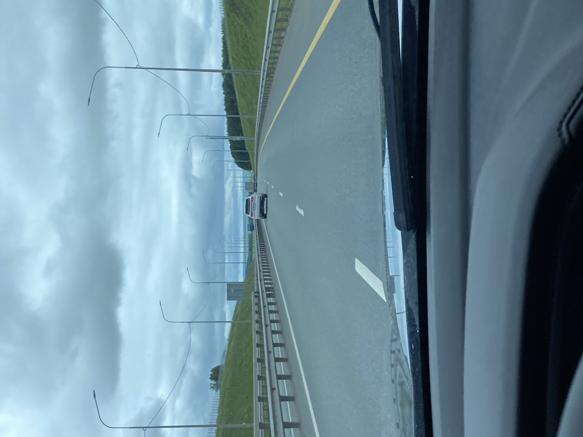 The image shows a view from inside a vehicle looking out onto a highway. The sky is overcast with heavy clouds. The highway has multiple lanes, separated by metal guardrails and white and yellow lane markings. Streetlights line the side of the road. On the right side of the highway, there is a grassy embankment. In the distance, there are a few vehicles, including a white SUV and a truck. The foreground of the image is dominated by the dashboard and windshield wipers of the vehicle. The image appears to be taken in a rural or suburban area, likely on a highway.
