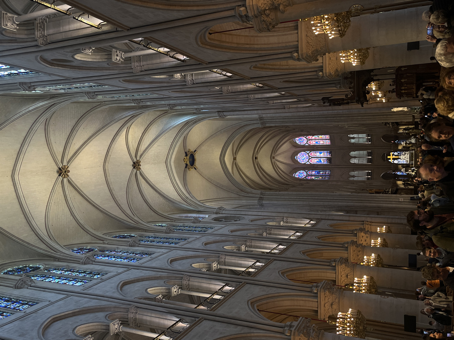 The image displays the magnificent interior of a large Gothic cathedral, viewed from a slightly elevated position looking down the nave towards the altar. The camera is rotated, presenting the elaborate vaulted ceiling as the main subject occupying the upper portion of the frame.
The cathedral's ceiling is characterized by intricate rib vaults in a light-colored stone, with ornate bosses at the intersections of the ribs. Along the upper walls, rows of large stained-glass windows, predominantly featuring shades of blue, filter natural light into the vast space. Below these, a triforium or clerestory level with arched openings runs along the nave. Massive stone columns line the nave, supporting the structure, and multiple ornate chandeliers with many lights hang from the ceiling, casting a warm glow.
In the foreground and to the right, a significant number of people are gathered. They appear to be visitors or congregants, many looking forward or upward, observing the grandeur of the architecture. Several individuals are seen holding up mobile phones, seemingly taking photographs. Their clothing is casual, typical of tourists.
Further down the nave, towards the end of the building, more vibrant stained-glass windows are visible, including several small rose windows and lancet windows with a richer palette of reds, blues, and yellows. A prominent, glowing golden cross stands near the far end, marking what appears to be the altar area. The illumination within the cathedral is a blend of bright natural light from the windows and the warm artificial light provided by the chandeliers and other fixtures, creating a dramatic and reverent ambiance. The architectural style, particularly the rib vaults and stained glass, strongly suggests a historic European cathedral, fitting with the location context of Notre Dame in Paris, France. No discernible text is visible in the image.