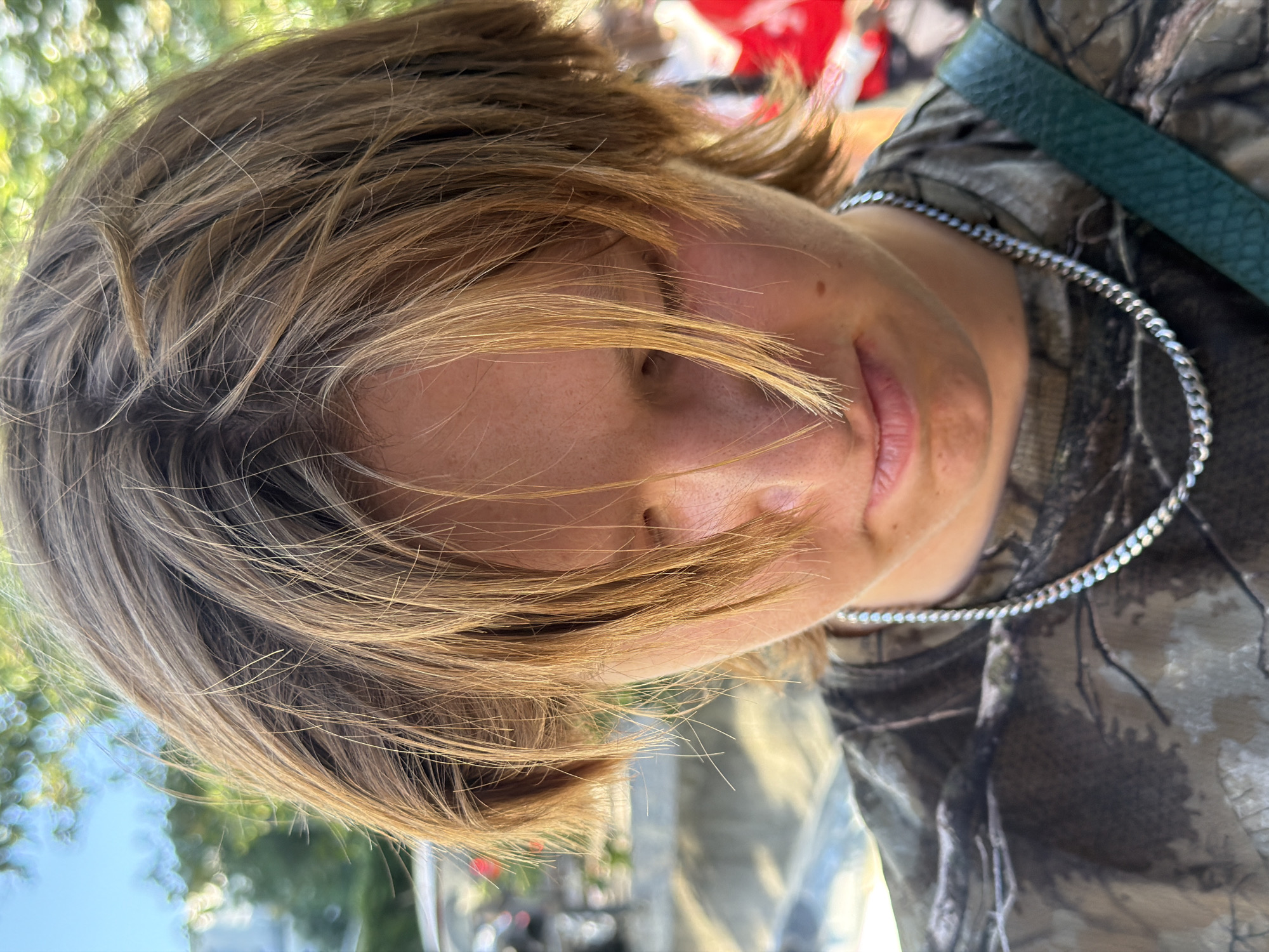 This is a close-up, head-and-shoulders portrait of a young person with light brown, shaggy hair falling over their face. They are wearing a camouflage t-shirt and a silver chain necklace. The background is out of focus, showing green foliage and a blue sky. There are hints of people and structures in the further background, suggesting an outdoor urban setting. The image does not provide any specific details to identify the location as Lyon, France.