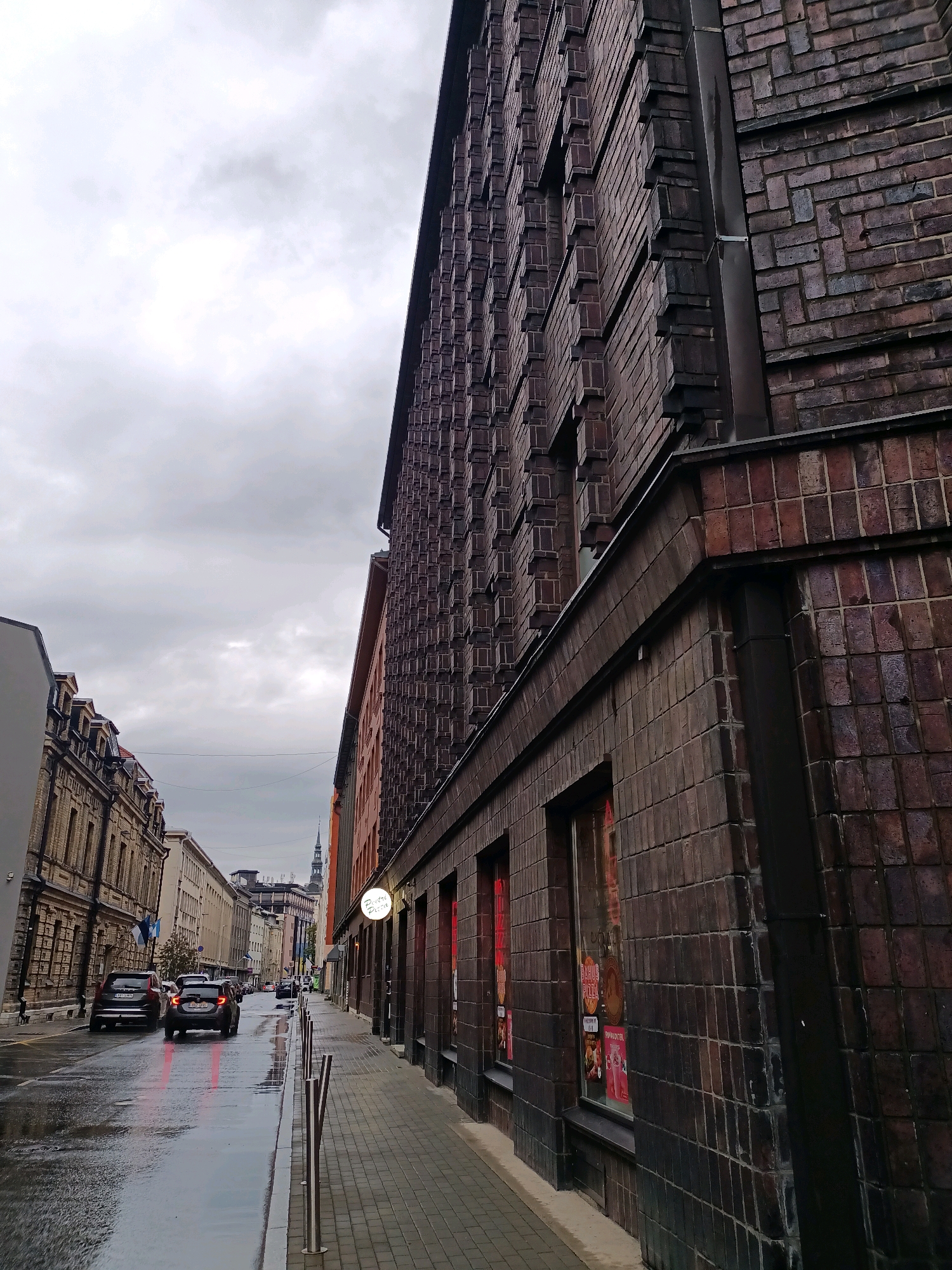 The image depicts a street scene in Tallinn, Estonia, likely during overcast or rainy weather, given the wet pavement and the gray sky. The perspective is from the sidewalk, looking down a city street flanked by buildings. On the right, a dark brick building occupies the foreground, its facade characterized by a patterned brickwork and multiple windows displaying red-lit advertisements. To the left of this building is a sidewalk with metal bollards. Across the street, to the left, are several other buildings. The first building on the left is a lighter-colored, multi-story structure with a detailed architectural style. Further down the street, other buildings with visible rooftops and spires can be seen. Two cars are driving on the wet street, their headlights and taillights illuminated. The overall impression is of an urban environment with historical architecture and active city life, albeit subdued by the weather.