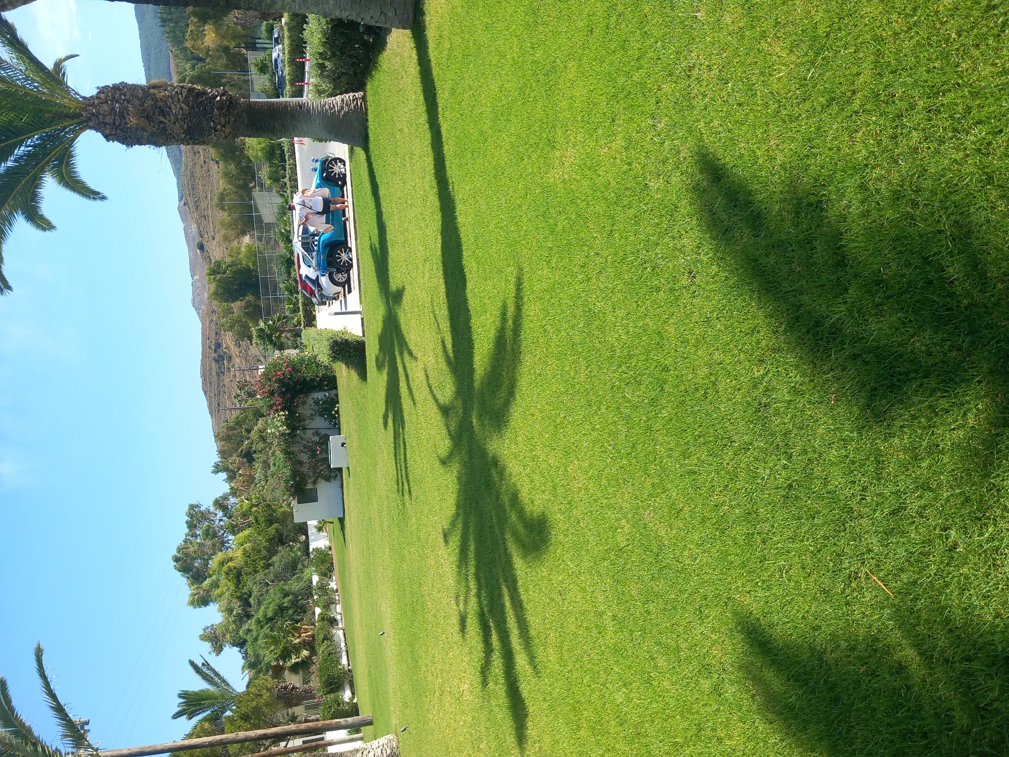 The image depicts a sunny outdoor scene with lush green grass in the foreground, a shadow of a palm tree cast across it. In the background, a blue buggy-like car is parked, with two people standing near it. Behind the car, there are several other parked vehicles, a low building with white walls, and dense vegetation including palm trees and flowering bushes. Further in the distance, hills covered in sparse vegetation rise under a clear blue sky. The scene appears to be a resort or a similar establishment in a warm climate, consistent with the city of Kos, Greece, known for its beaches and palm-lined streets.