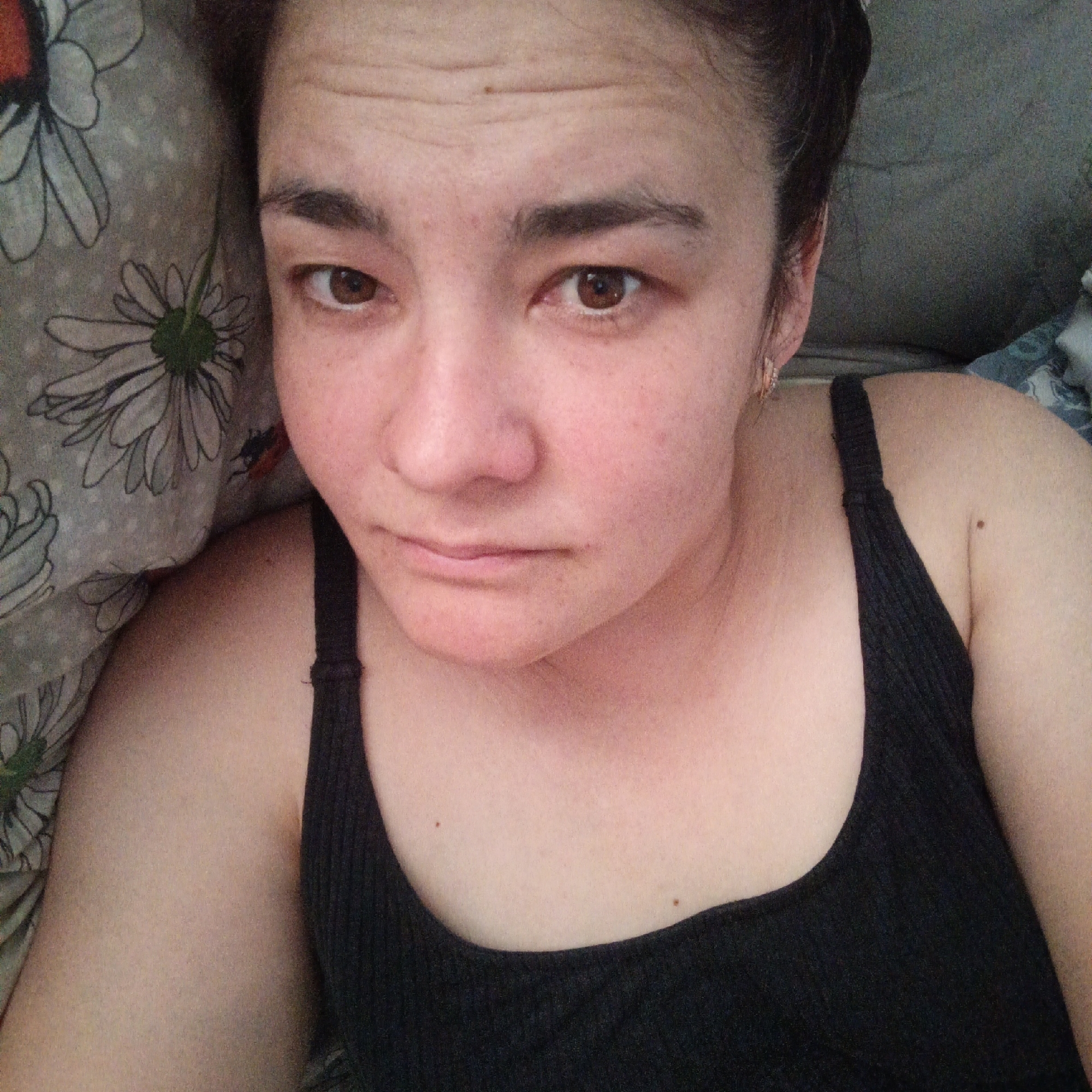 This is a selfie of a woman lying down, looking at the camera. She is wearing a black tank top and has brown eyes and dark hair pulled back. Her skin is fair with some freckles and redness. She appears to be indoors, possibly in a bedroom, as suggested by the floral patterned fabric behind her and the gray background. There is no indication of the location being Podlubovo, Russia, or any other specific city. The image is a close-up, focusing on her face and upper body.