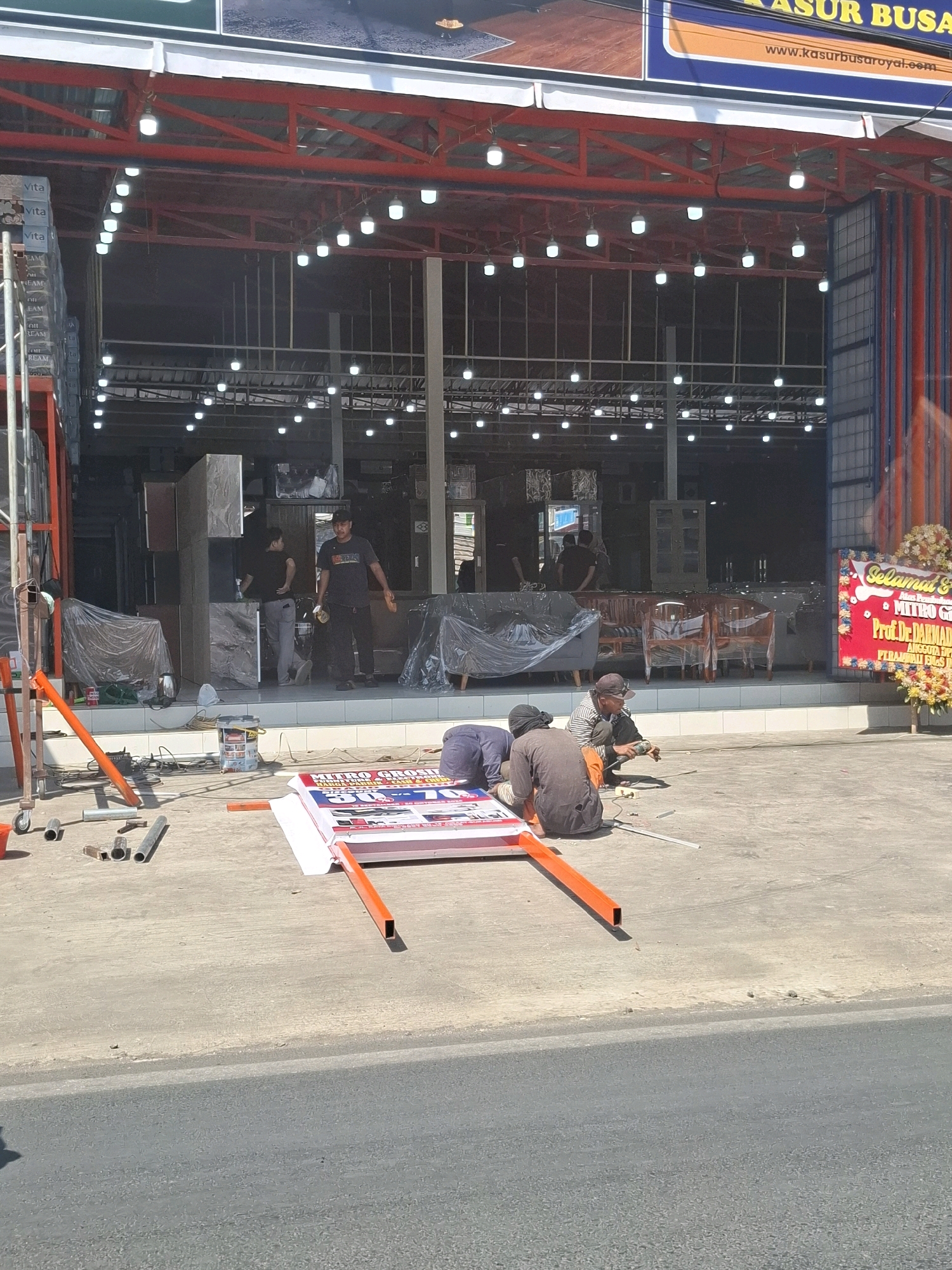 The image depicts the exterior of a store located in Selajambe, Indonesia, specializing in mattresses and furniture. The storefront features a red metal frame structure with numerous lights illuminating the interior. A banner with Indonesian text and graphics advertising discounts is prominently displayed on the ground in the foreground, supported by orange metal poles. Several individuals are present, appearing to be workers engaged in setting up or dismantling the banner. One person is actively working on the banner with a tool, while others are positioned around it. In the background, the store interior reveals stacked mattresses, furniture covered in plastic sheeting, and additional merchandise. The overall scene suggests an ongoing commercial activity or preparation for an event at the establishment.