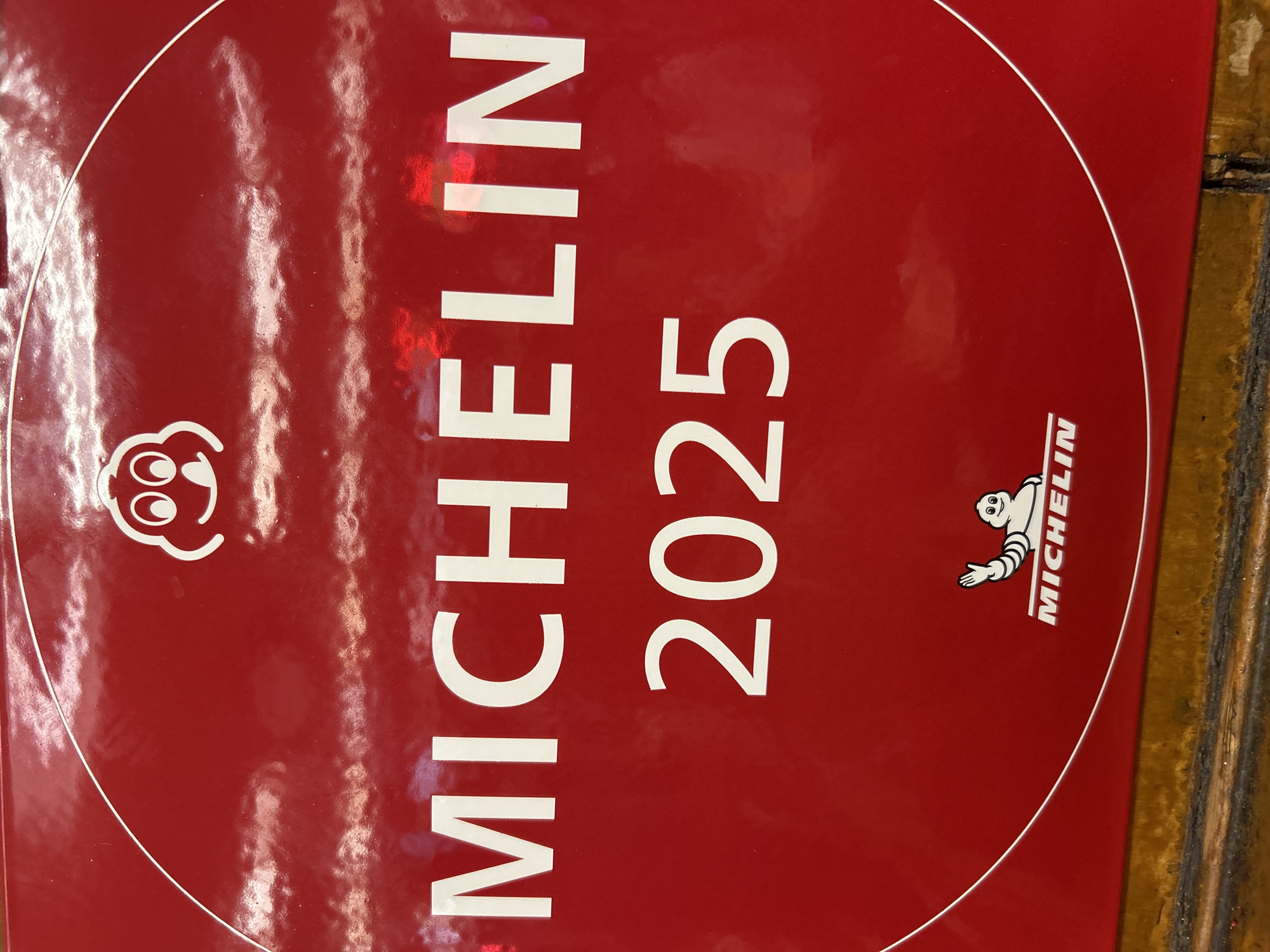 The image shows a Michelin Guide 2025 publication cover. It's red, with "MICHELIN 2025" printed in white. The Michelin Man logo is in the lower right corner. There is a head in the upper left corner. The cover appears to be glossy, with some surface reflections visible. It is sitting on a brown and black surface, possibly wooden. Without further information, there is no indication of its location in New York City, USA.