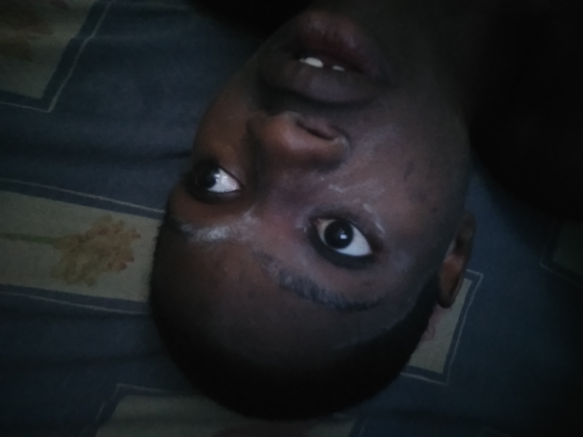 An individual with dark skin lies on a patterned surface. The head is tilted upward and to the side, with eyes open and mouth slightly ajar. Visible facial features include the nose, lips, and eyebrow hair. The individual appears to be in a dark environment. The surface beneath them shows a repeating geometric pattern with floral accents. The location is not discernable from the image.