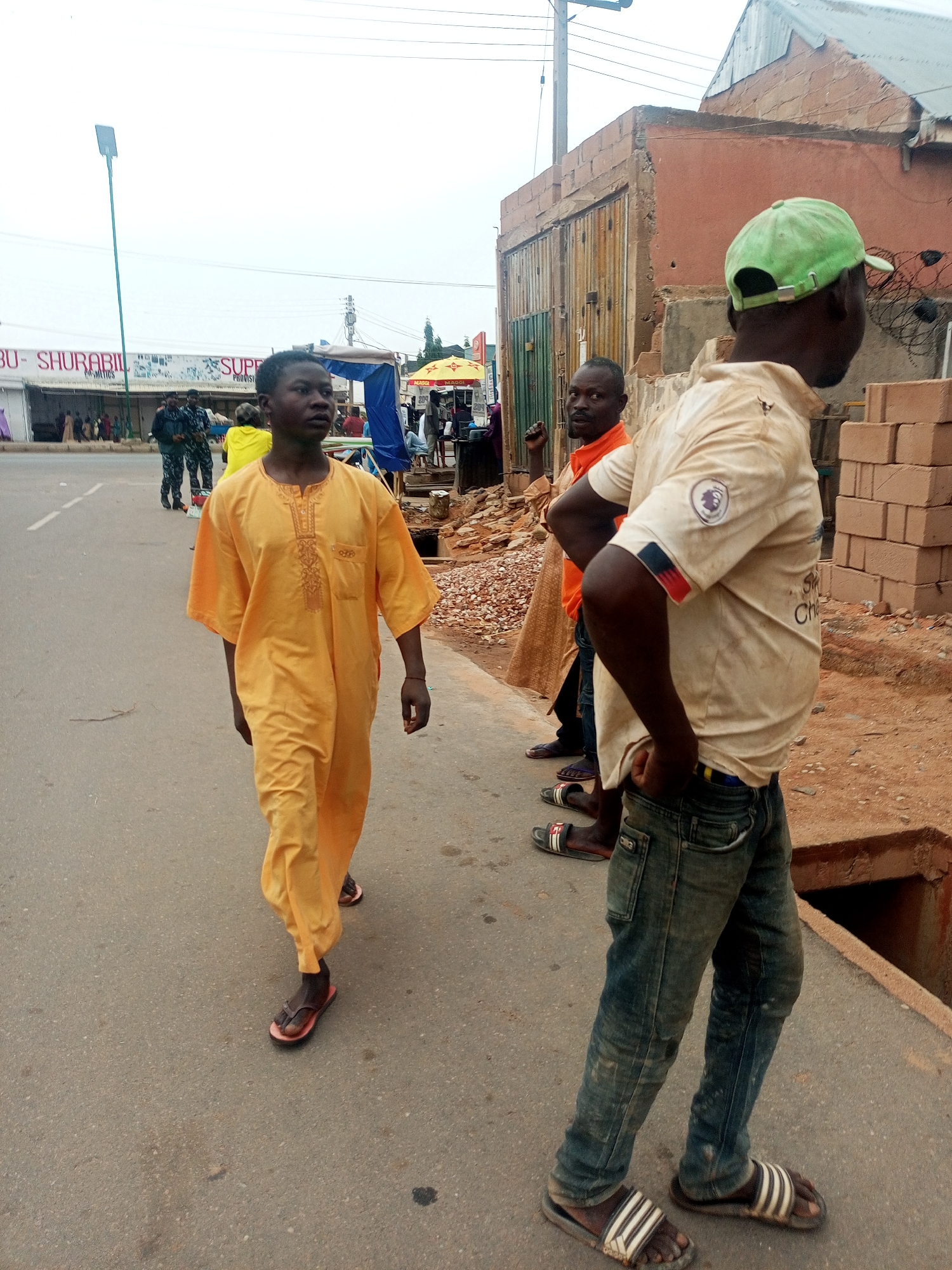 The image shows a street scene in what is described as the city of Gombe, Nigeria. In the foreground, a young man wearing a long, gold-colored robe and flip-flops walks toward the camera along a paved street. To the right, another man in a light-colored shirt, jeans, and a green baseball cap stands with his hand in his pocket. Behind him and slightly to the left is a third man wearing an orange shirt. In the background, a building with a metallic gate is visible. Further back across the street are buildings and structures with signs, and people milling about. Two uniformed officers can be seen standing near a shop. The sky appears overcast. There is exposed dirt on the sidewalk, and stacks of red bricks are near the standing man.