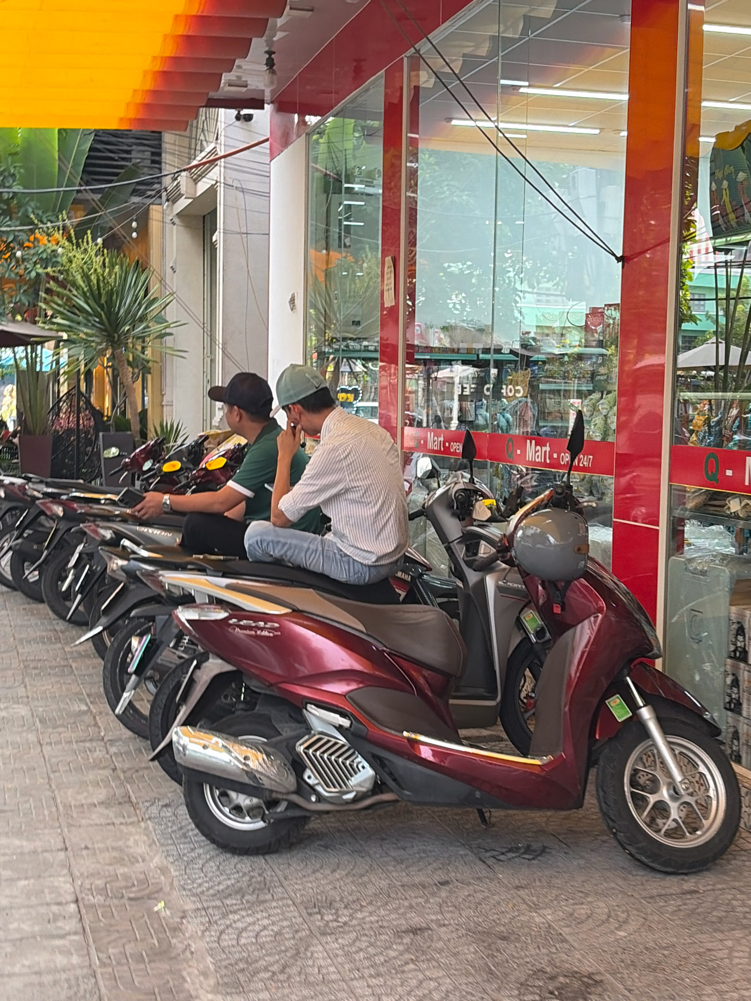 This is a medium shot of two men sitting on motorcycles parked on a sidewalk in front of a convenience store in Đà Nẵng, Vietnam. The store, identified as "Q-Mart," appears to be open 24/7. The men are both wearing casual clothing. The man on the left is wearing a dark green polo shirt and a black baseball cap, while the man on the right is wearing a striped button-down shirt and a light green cap. They are both looking towards the store. Several other motorcycles are parked in a line behind them, suggesting a common mode of transportation in the area. The sidewalk is paved with textured tiles. The background shows the storefront with large glass windows reflecting the surroundings, including trees and buildings, and a brightly colored awning overhead. The overall scene depicts a typical urban setting in Southeast Asia.