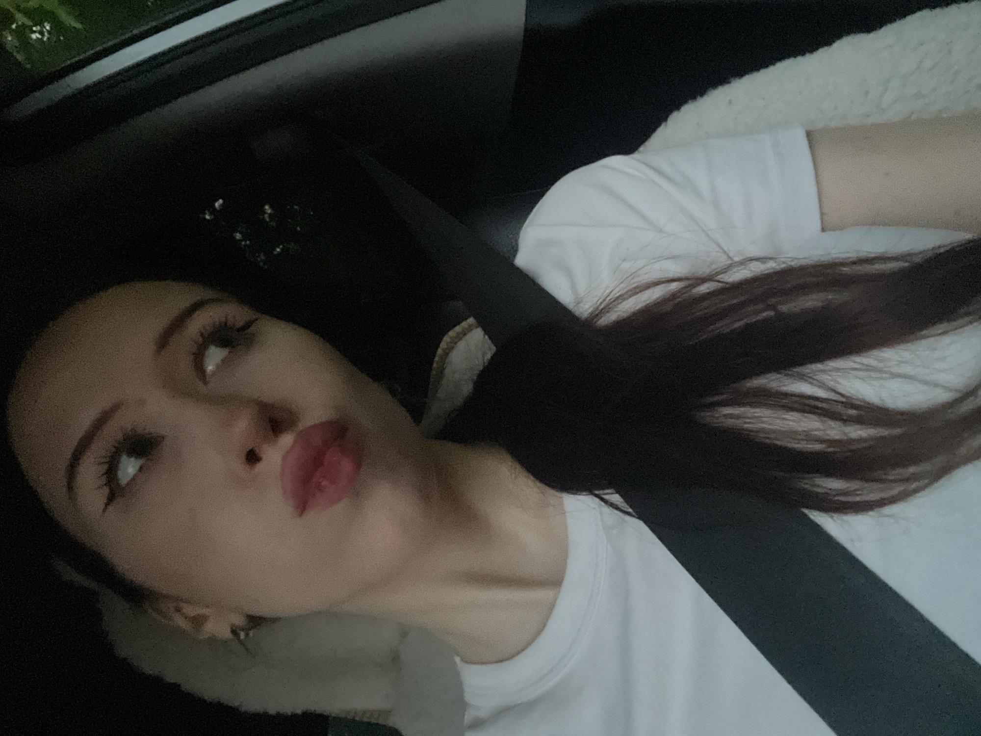 A young woman with dark hair, wearing a white t-shirt and a seatbelt, is in a car. She is looking to her right with her lips pursed, and her right hand is not visible. The environment appears to be outdoors, with trees visible in the background.