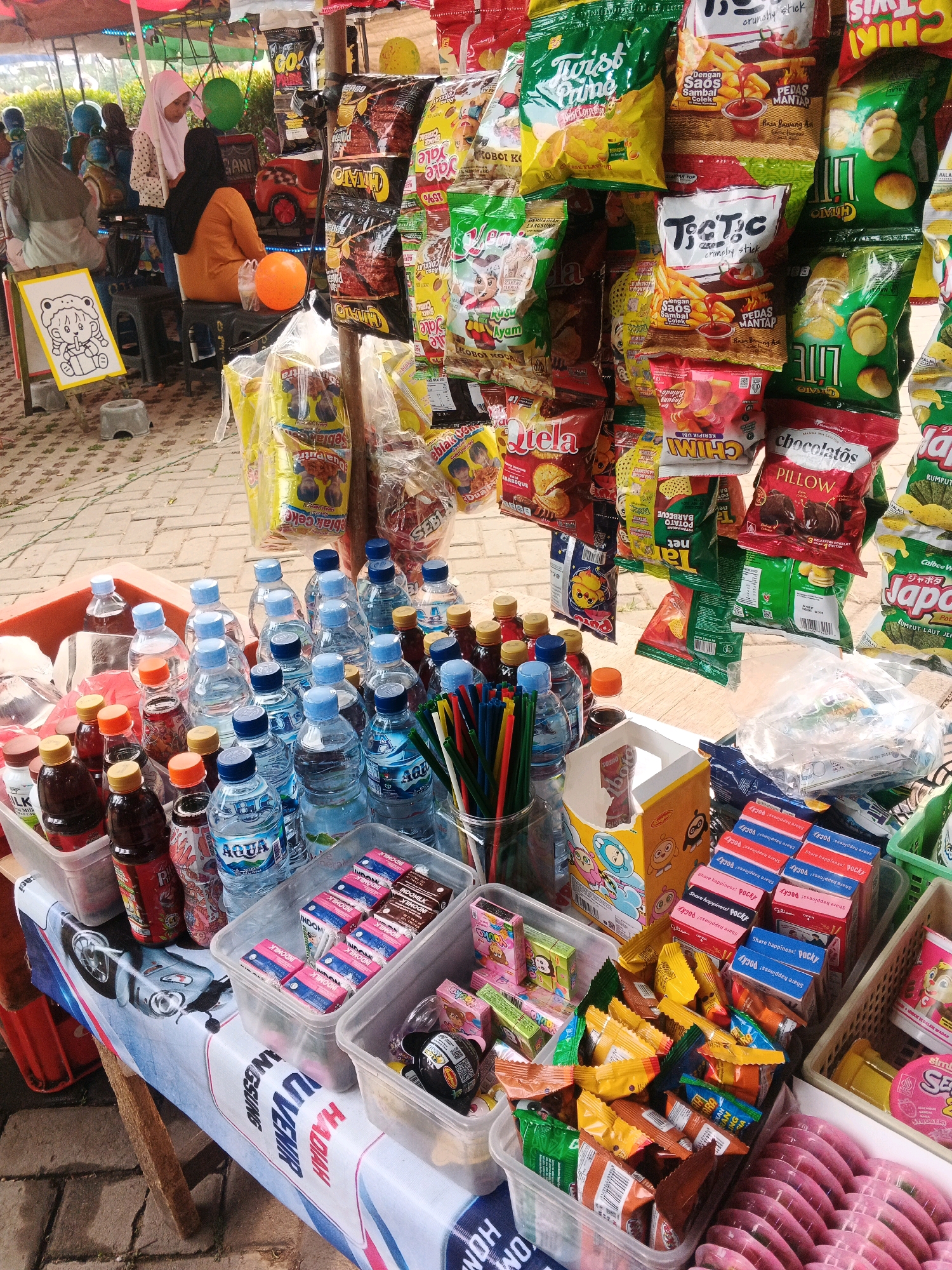 The image depicts an outdoor market stall in Curug Kulon, Indonesia, selling a variety of snacks and drinks. The stall is set up with several shelves and containers displaying an array of colorful packaged goods, including chips, cookies, and bottled beverages. In the foreground, plastic containers hold bottled water and other drinks, alongside smaller packaged snacks. A table covered with a patterned cloth, featuring an image of a scooter and text in Indonesian, is laden with more products. In the background, several individuals, including women wearing headscarves, are visible, some interacting with the stall or observing the surroundings. The ground is paved with bricks, and the overall setting suggests a casual, community-oriented marketplace.