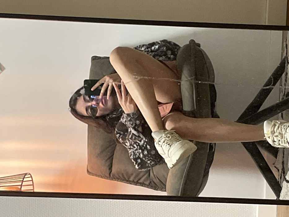 A woman in sunglasses sits in a cushioned chair, taking a selfie with her phone. She wears a patterned jacket and shorts. Her legs are propped up on the armrest of the chair, with sneakers on her feet. The image is a reflection in a mirror, revealing part of a radiator, a black metal structure, and a white wall in the background.