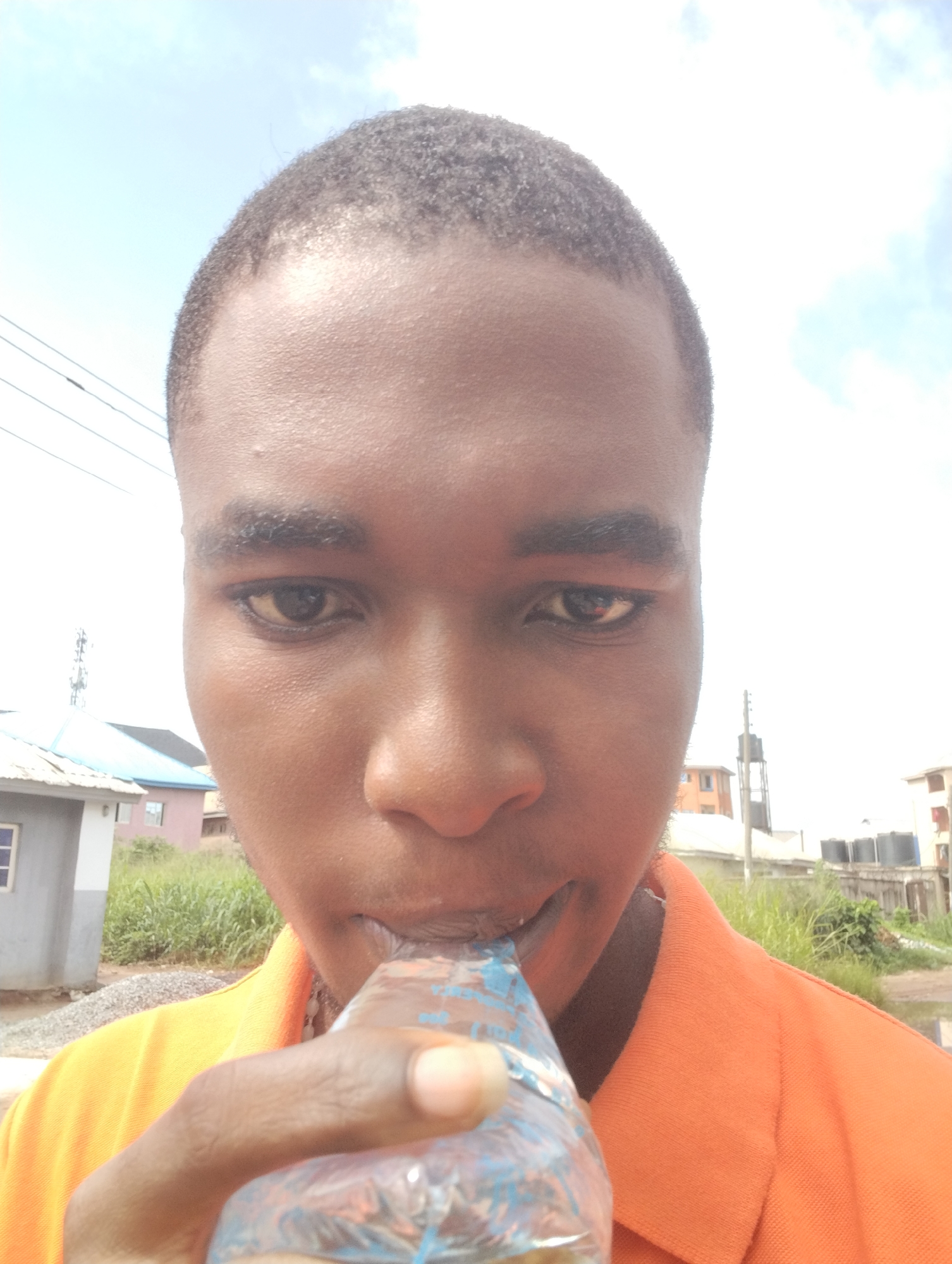 A close-up, slightly low-angle portrait captures a young Black man outdoors in Ihiagwa, Nigeria. He occupies the central and lower portions of the frame, with his face prominent. He has short, dark, textured hair and dark eyes. He is wearing an orange collared shirt.
The primary activity depicted is the man drinking from a small, transparent plastic sachet or bottle, which he holds to his mouth with his right hand. The sachet has indistinct blue markings. His expression is neutral as he looks directly at the camera, seemingly concentrating on drinking.
The background suggests an outdoor, possibly residential or semi-rural setting under bright daylight. A light blue sky with scattered white clouds fills the upper half of the image. To the left, utility wires and a pole are visible, along with parts of a building featuring a light-colored roof (possibly corrugated iron) and blue accents. On the right, another building and a tall structure with what appears to be a water tank on top are seen in the distance. The ground immediately behind the man features green grass and other vegetation, and an unpaved area of dirt or gravel can be seen on the left. The overall impression is a sunny day in a developing area. No legible text is visible in the image.