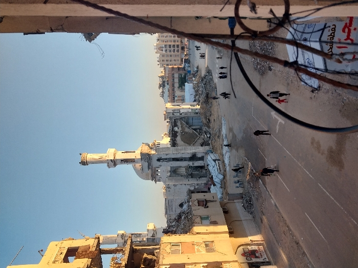 The image depicts a devastated urban street scene in Khan Yunis, Palestinian Territory, under a clear, bright sky. The central feature is a heavily damaged mosque with its minaret largely intact but the main structure partially collapsed, surrounded by rubble. To its left, a multi-story building is severely compromised, showing exposed structural elements and large sections missing. Further left, another building, while damaged, exhibits a partially functional ground floor with a visible storefront.
The street, traversing from the foreground towards the mosque, is a paved road marked with white lines and a directional arrow, but it is partially covered with dirt and debris. Piles of concrete and building fragments are visible along both sides of the street.
Several individuals are walking on the street. In the lower-right foreground, an adult male holding the hand of a small child in a red garment walks towards the background. Other individuals, singly and in small groups, are scattered across the street and closer to the mosque. On the left side of the street, near a damaged building, a small cluster of plastic chairs is visible. The immediate foreground on the right is framed by bent metal bars and exposed rebar from a damaged structure, with partially visible Arabic signage below, including the words "نقطة شحن" (charging point) and "شكرا" (thank you). Buildings in the background also display varying degrees of damage.