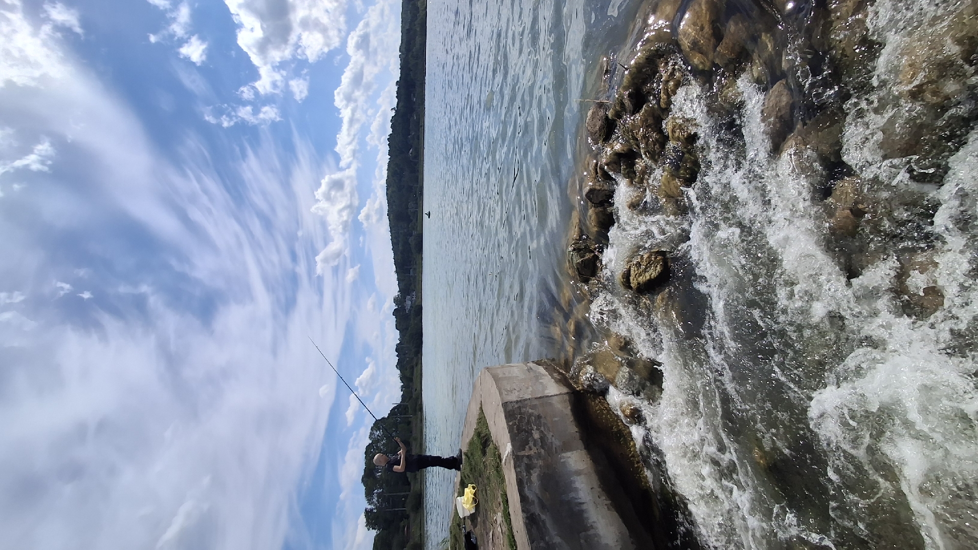 This vertical image captures a daytime scene of a person fishing from a concrete structure in Krasnoye Selo, Russia. A young male individual, wearing a dark short-sleeved top and dark trousers, stands on the light-colored concrete platform, holding a long fishing rod with the line cast into the water. A yellow item, possibly a bag, is visible on the concrete to his left. Immediately below and to the right of the platform, water churns and froths as it flows rapidly over a collection of visible rocks and stones. The main body of water, extending into the mid-ground, appears calm and has a light greenish-blue hue. In the distant background, a low, tree-covered hill defines the horizon, with faint structures discernible on the left side. A small, dark, oblong object floats on the water in the far distance. The sky, occupying the upper portion of the frame, is blue with numerous white clouds, ranging from wispy cirrus to more defined cumulus formations.