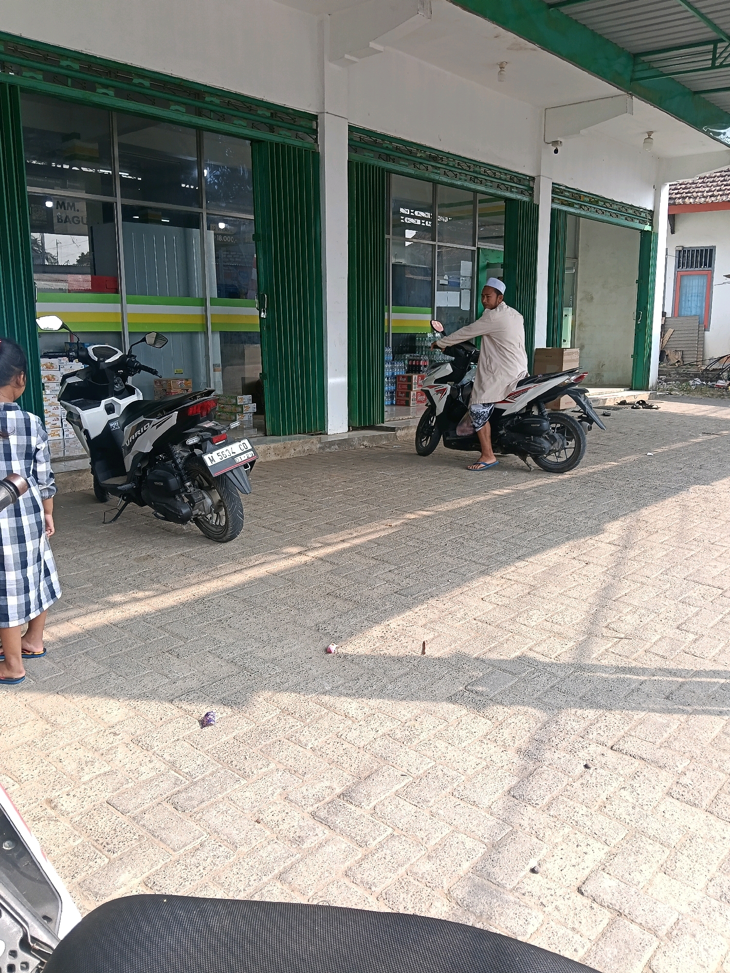 A person wearing a white robe and cap is sitting on a white and black motorcycle. The motorcycle has a license plate that reads "M 5634 CD". The person is looking towards the left. To the left of the motorcycle, a young child wearing a blue and white plaid dress and flip-flops is standing. In the background, there is a building with green metal roll-up doors. The building appears to be a store. The ground is paved with interlocking bricks. Sunlight is casting shadows on the ground.