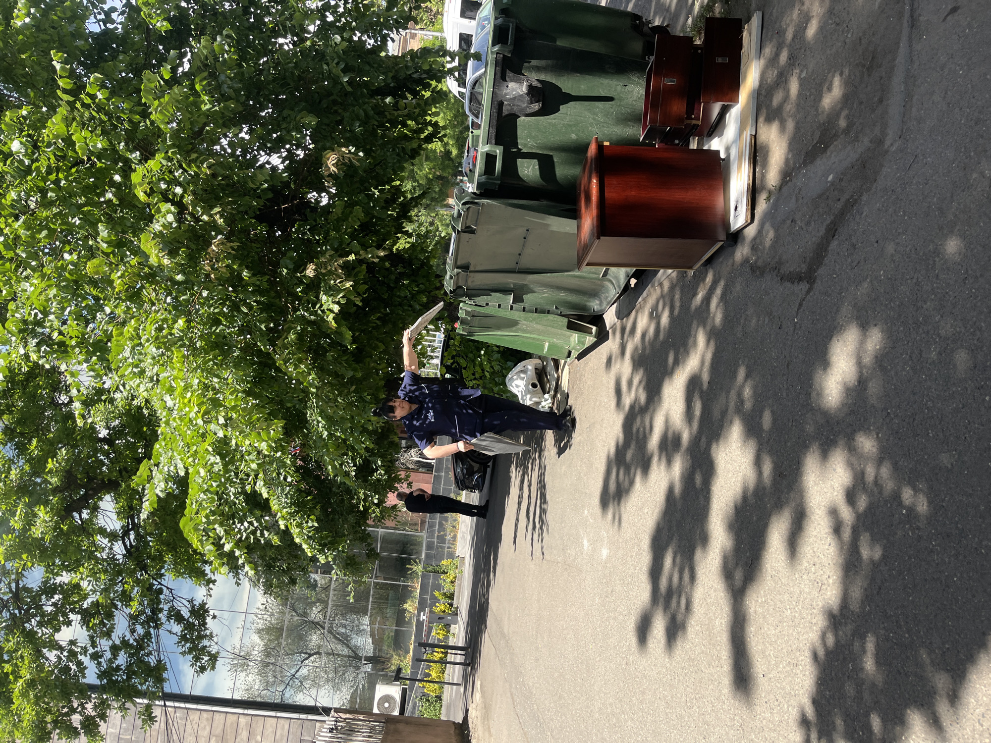 A daytime, angled shot of a city street, likely in Yerevan, Armenia. The main subject is a person in a blue uniform standing under a tree, appearing to be sorting through materials, possibly papers, near several green trash bins. To the right of the person are two dark wood cabinets atop a white platform. Another person, dressed in black, stands in the background near a metal fence and a green, greenhouse-like structure. The area is paved, with shadows of trees cast on the ground. The photo gives a glimpse of urban life with waste management and possible discarded furniture.