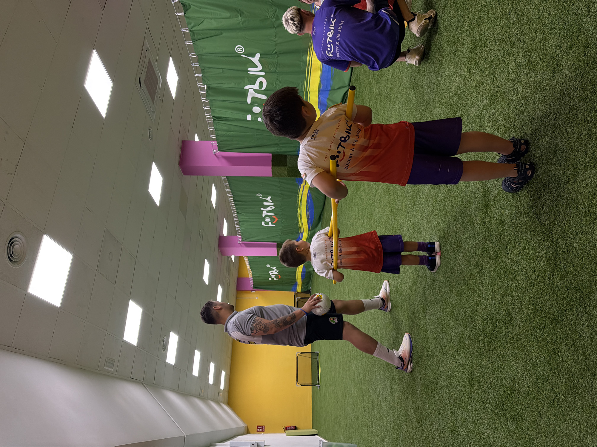 This media file depicts an indoor setting with artificial turf, likely a sports facility. The scene features three individuals: an adult male and two young boys. The adult, wearing a grey t-shirt and black shorts, is holding a soccer ball. One of the boys, in a white and orange t-shirt and purple shorts, is holding a yellow stick. The other boy, in a purple t-shirt, is partially visible in the background.
The individuals appear to be engaged in a soccer-related activity or training session. The backdrop includes green banners with repeating text. Visible text on the banners includes "FITBIK," "10TH ANNIVERSARY," and "soccer & life skills," suggesting the name of a sports academy or event. The lighting suggests an indoor environment, possibly during the daytime. The overall atmosphere seems to be focused and instructive. The location appears to be a dedicated space for sports training with a padded wall and bright lighting from the ceiling.