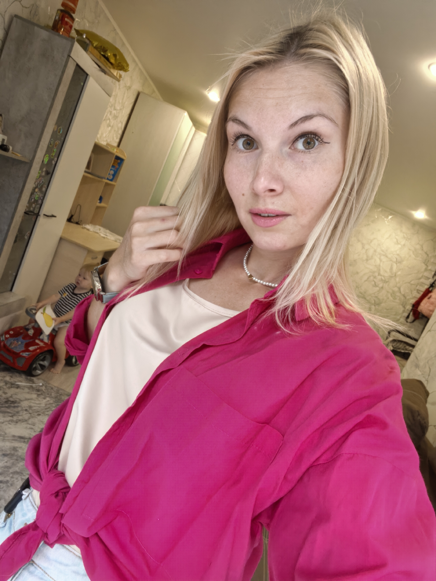 A young woman with blonde hair and light brown eyes is taking a selfie in a domestic setting. She is wearing a bright pink button-down shirt, tied at the waist, over a white tank top and light-colored jeans. A pearl necklace with a small gold charm is visible around her neck. In the background, a toddler is sitting on a red toy car, facing away from the camera, wearing a striped t-shirt and diaper. The environment appears to be a living room or playroom, with furniture, shelves, and a wall with textured wallpaper. The overall lighting suggests indoor illumination. The location is identified as Sterlitamak, Russia.
