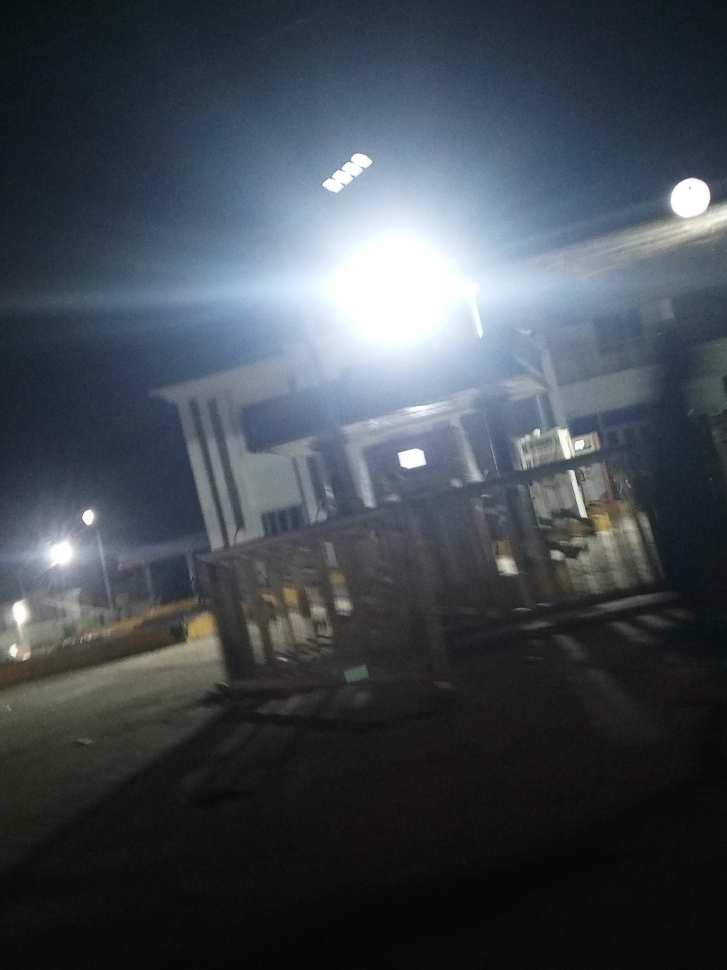 The image depicts an outdoor scene at night. The location appears to be a construction site or an area undergoing renovation, based on the presence of wooden structures and scaffolding-like elements. The buildings in the background have a modern architectural style with visible columns. The scene is brightly lit by overhead lights. The ground appears to be paved.