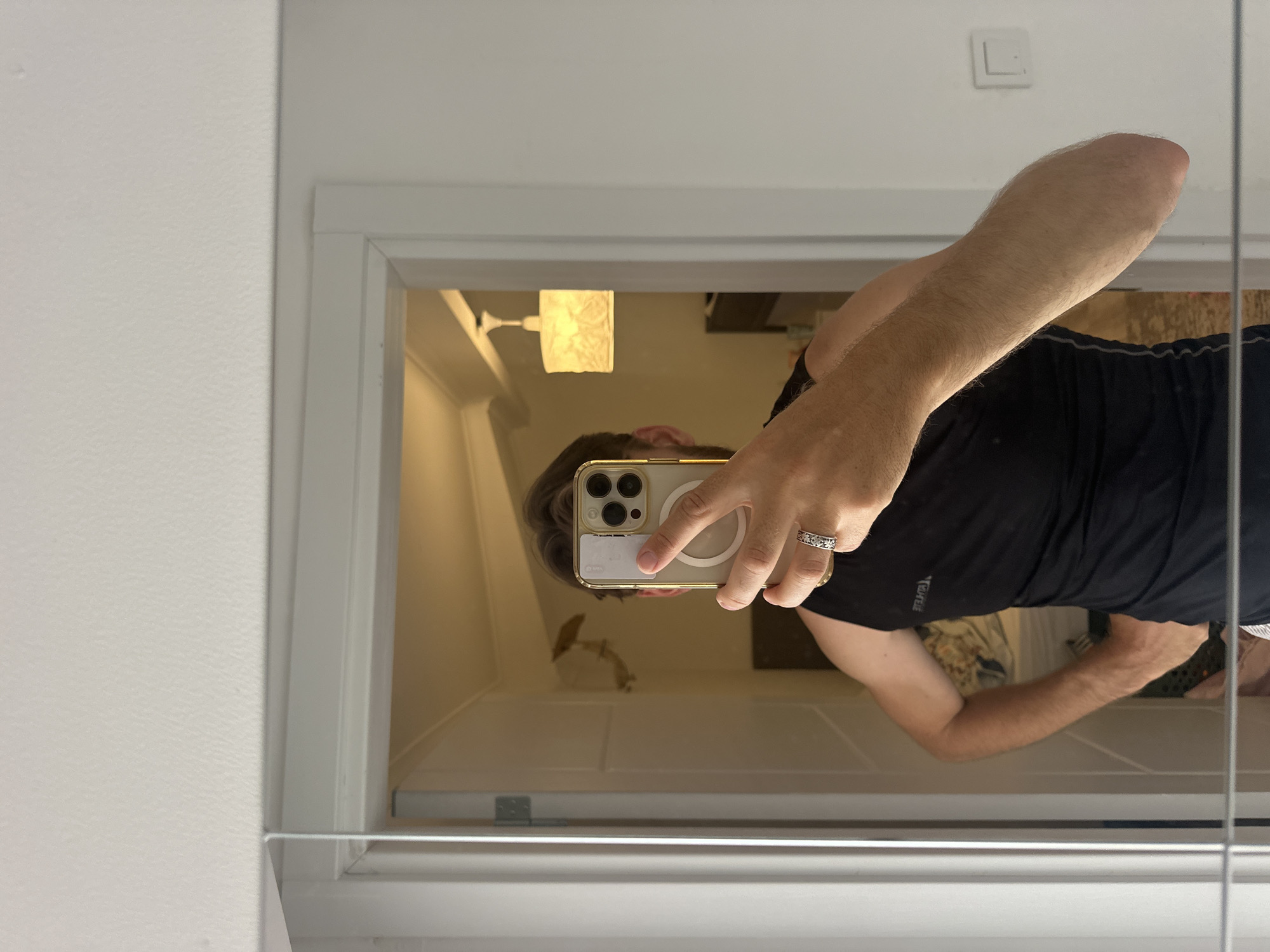 A person takes a mirror selfie with a gold iPhone. The reflection shows a bathroom interior with a light fixture and a partial view of a bedroom. The location appears to be indoors, but there is no clear indication of Fethiye, Turkey.
