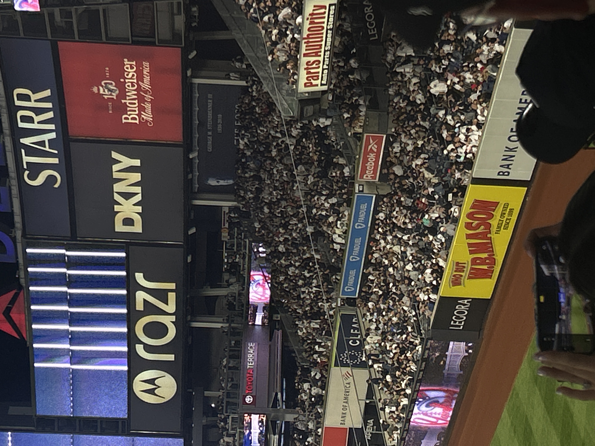 This image captures an event at Yankee Stadium in New York City. A densely packed crowd fills the stadium seating. In the foreground, a person is holding up a smartphone to take a picture. Above the crowd, there is a series of advertisements for various brands, including Starr, DKNY, Budweiser, Motorola Razr, Toyota Terrace, Bank of America, Clear, Fanduel, Reebok, Parts Authority, and W.B. Mason. A memorial for George M. Steinbrenner can also be seen.