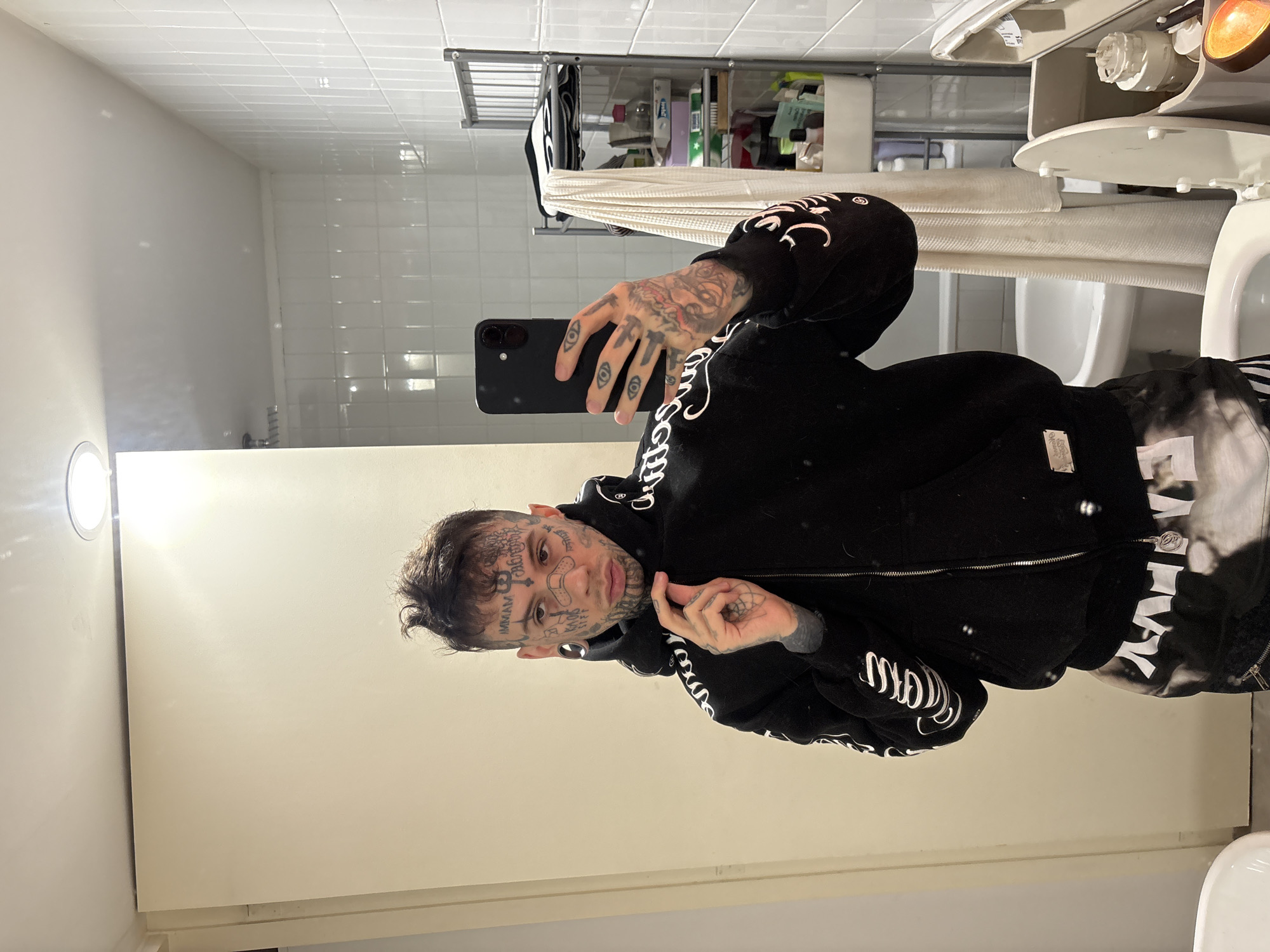 A person with numerous facial tattoos and tattooed hands is taking a selfie in a bathroom. The person is wearing a black zip-up hoodie with white writing on the sleeves. The bathroom has white tiled walls and a mirror. A toilet and a metal shelving unit holding toiletries are visible in the background. The location is unknown and there is no visible evidence linking the image to Buenos Aires, Argentina.
