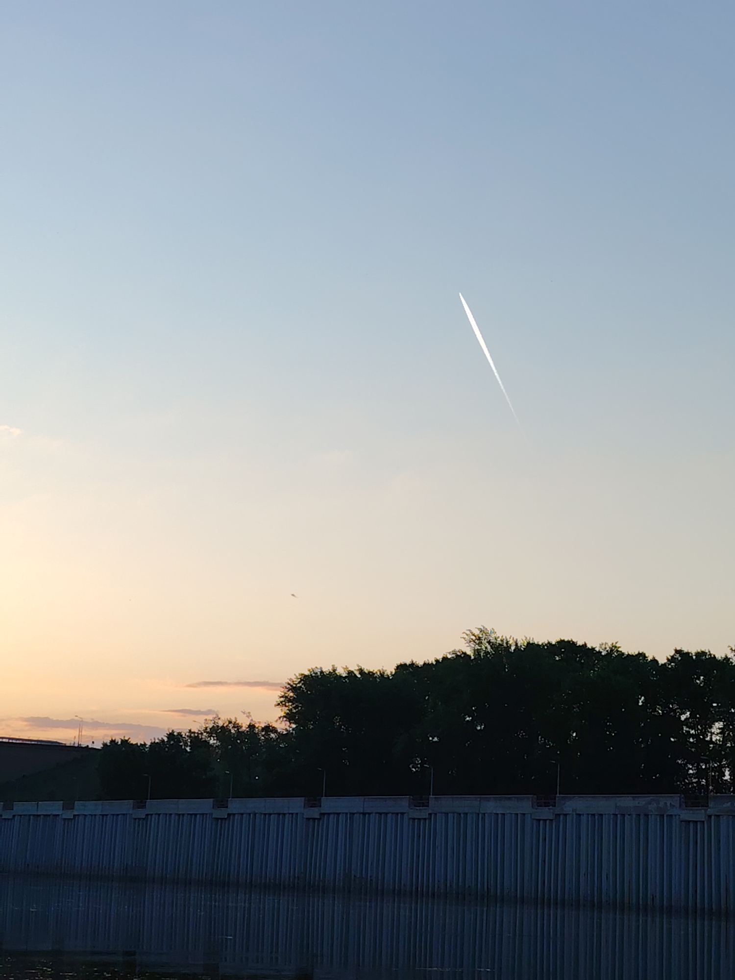 The image shows a riverside scene in Chaikovsky, Russia, taken during a sunset. The sky is a gradient of light blue to orange. A contrail from a jet is visible in the sky. Below the sky is a line of trees, and in the foreground, a concrete embankment is visible next to a body of water. The water reflects the patterns of the embankment.