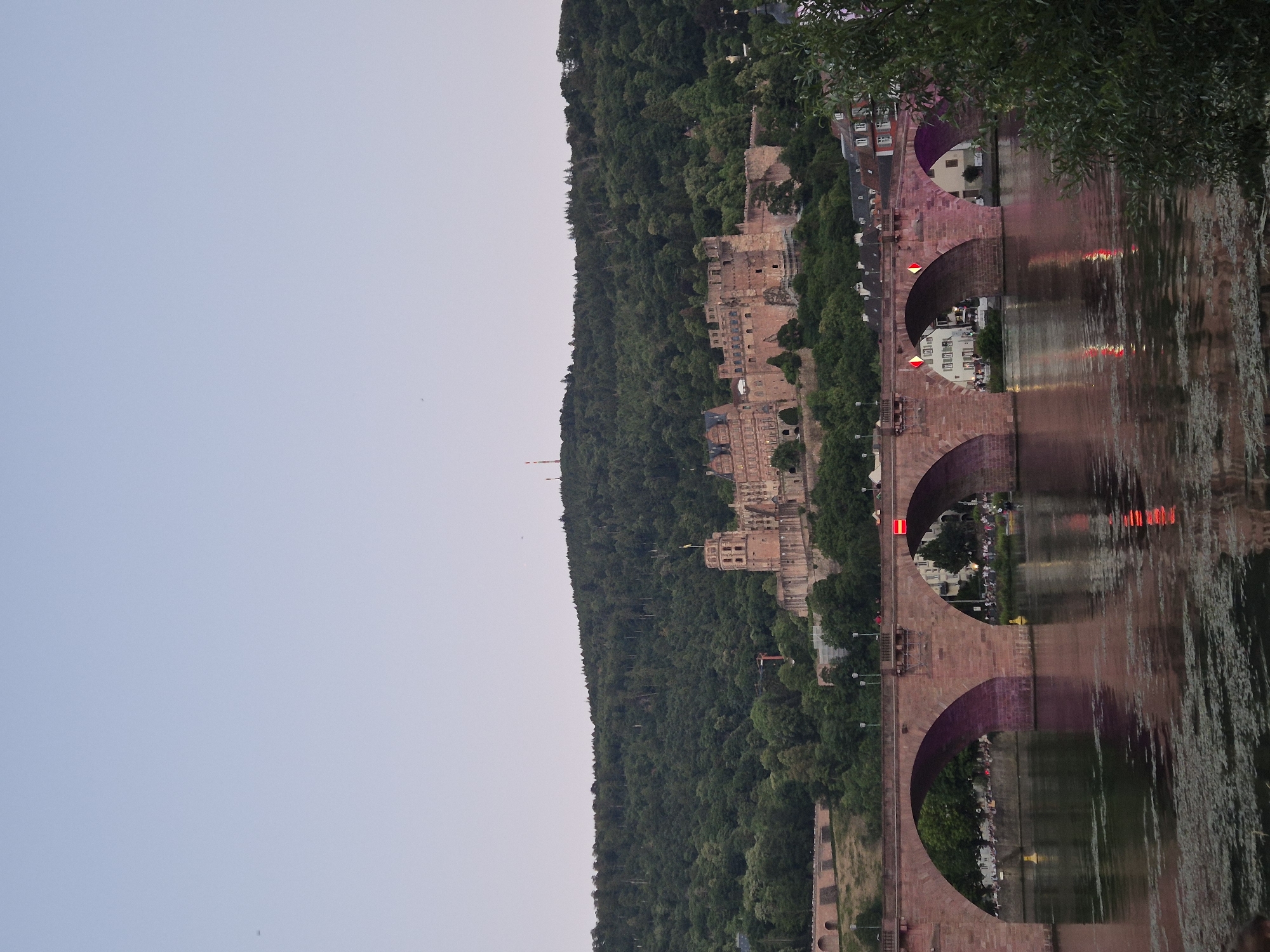 This image depicts a scenic view of Heidelberg Castle and the Old Bridge (Alte Brücke) over the Neckar River at dusk. The castle, a large, historic structure with stone towers and walls, is perched on a wooded hillside. The Old Bridge, made of stone with multiple arches, spans the river, and its reflection is visible in the water. The river is calm, with ripples on its surface. A lush green forest covers the hillside behind the castle. The sky is a pale, gradient blue, transitioning to a lighter hue towards the horizon. There are some lights on the bridge, casting a pinkish glow. A few people are visible on the bridge and along the riverbank. Trees and some buildings are present in the foreground and background, contributing to the overall picturesque setting.