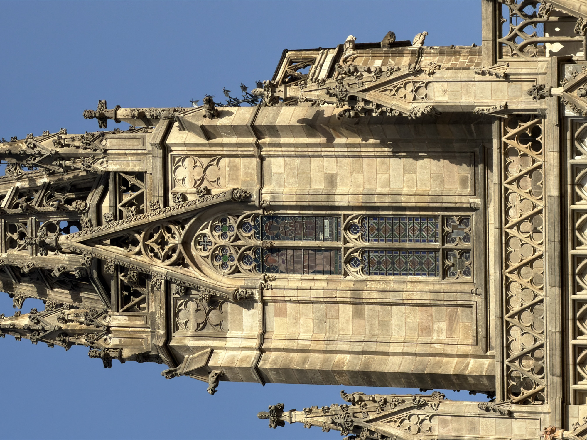 This image shows a detailed view of the bell tower of the Barcelona Cathedral, a Gothic Roman Catholic cathedral located in Barcelona, Spain. The tower is constructed of light-colored stone blocks and is adorned with intricate Gothic architectural elements. These include pointed arches, tracery, gargoyles, and decorative carvings. A prominent feature is a tall, narrow window with stained glass depicting religious figures. The blue sky serves as a backdrop, highlighting the architectural details of the tower. The lighting suggests it is daytime, with sunlight casting shadows on the stonework.