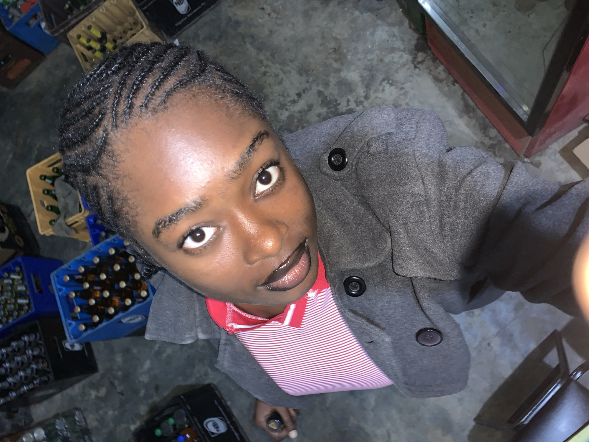 The image displays a person, likely female, looking up directly at the camera from a low angle. She has dark skin and her hair is styled in neat cornrows. Her expression is neutral to slightly smiling, with her mouth slightly open and glossy lips. She is wearing a dark grey jacket with three visible black buttons over a red and white striped polo shirt.
The background indicates an indoor commercial setting, possibly a beverage store or a storage room. Numerous crates filled with glass bottles are stacked around her; some are blue plastic, others are brown or black. A glass-fronted refrigerator or display unit is partially visible on the right side of the frame. The floor appears to be a dark, rough concrete surface. In the bottom center, a hand is visible holding a small, dark bottle.
The lighting is artificial, suggesting an indoor environment where the time of day is not clearly indicated. The scene is consistent with a commercial establishment, potentially in a location like New Stadium, Nigeria, where drinks are sold or stored. No distinct text or branding is clearly legible on any of the crates, though some faint markings can be observed. The subject's direct gaze implies an engaged pose for the photograph.