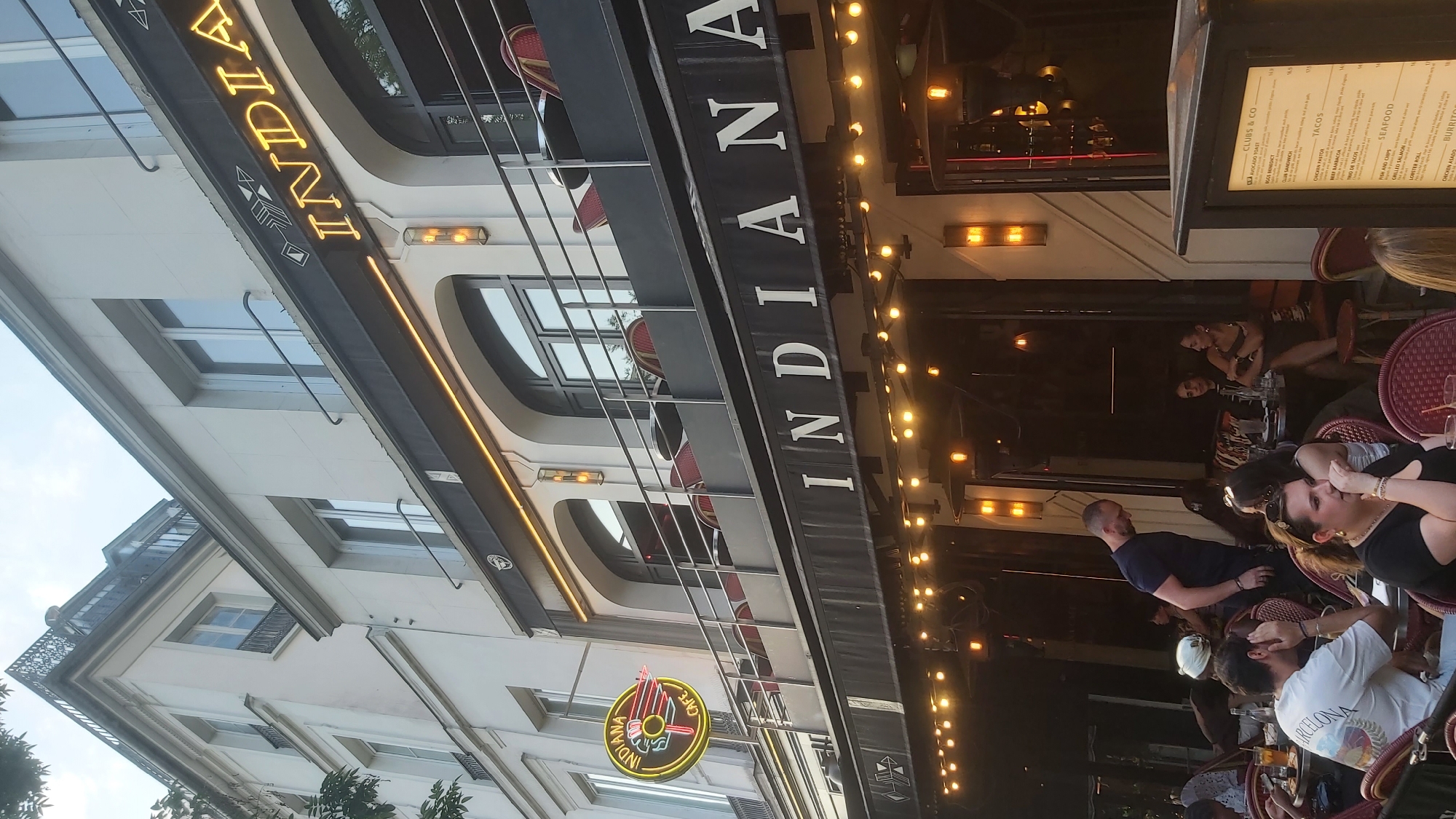 The image shows a multi-story building with the sign "INDIANA CAFE" prominently displayed on the upper floor and "INDIANA" on the awning of the ground floor. The building has a classical architectural style with multiple windows and balconies. The ground floor appears to be a restaurant or cafe with outdoor seating, illuminated by string lights. Several patrons are seated at tables, some engaged in conversation or eating. A menu is visible on a stand to the right. The overall scene suggests a lively urban environment, likely in a European city given the architectural style. The sky is visible at the top of the image, suggesting it is daytime.