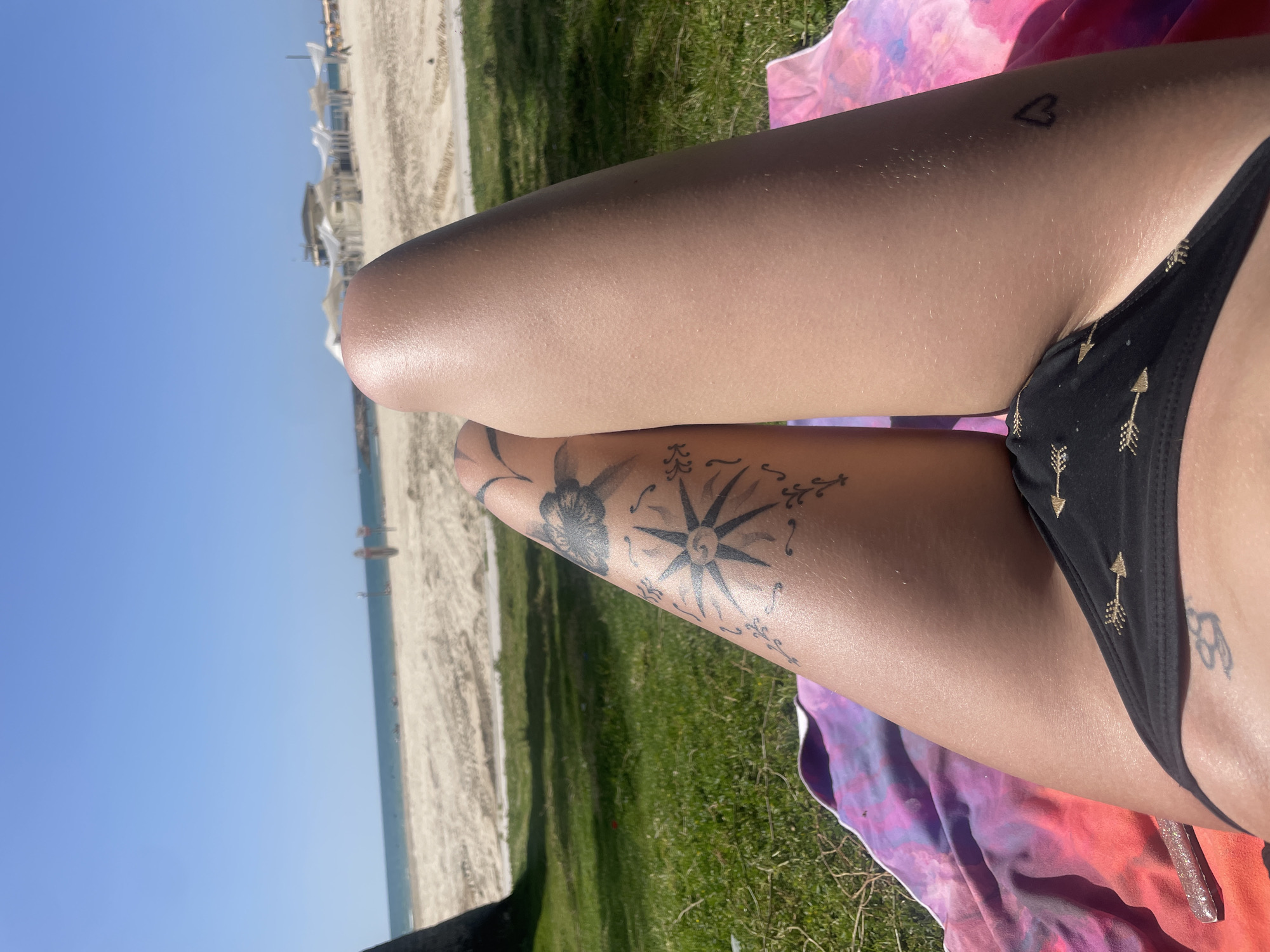The image depicts a woman lying on a multicolored blanket on grass near a beach. She is wearing a black bikini with gold arrow patterns. Tattoos are visible on her legs, including a compass rose, a floral design, and a small heart. In the background, there is a sandy beach, the sea, and a clear blue sky. Structures resembling beach umbrellas and small buildings are visible on the beach. Based on the context and visual cues, the location appears to be a beach in Herzliya, Israel, which is known for its coastal areas and beaches.