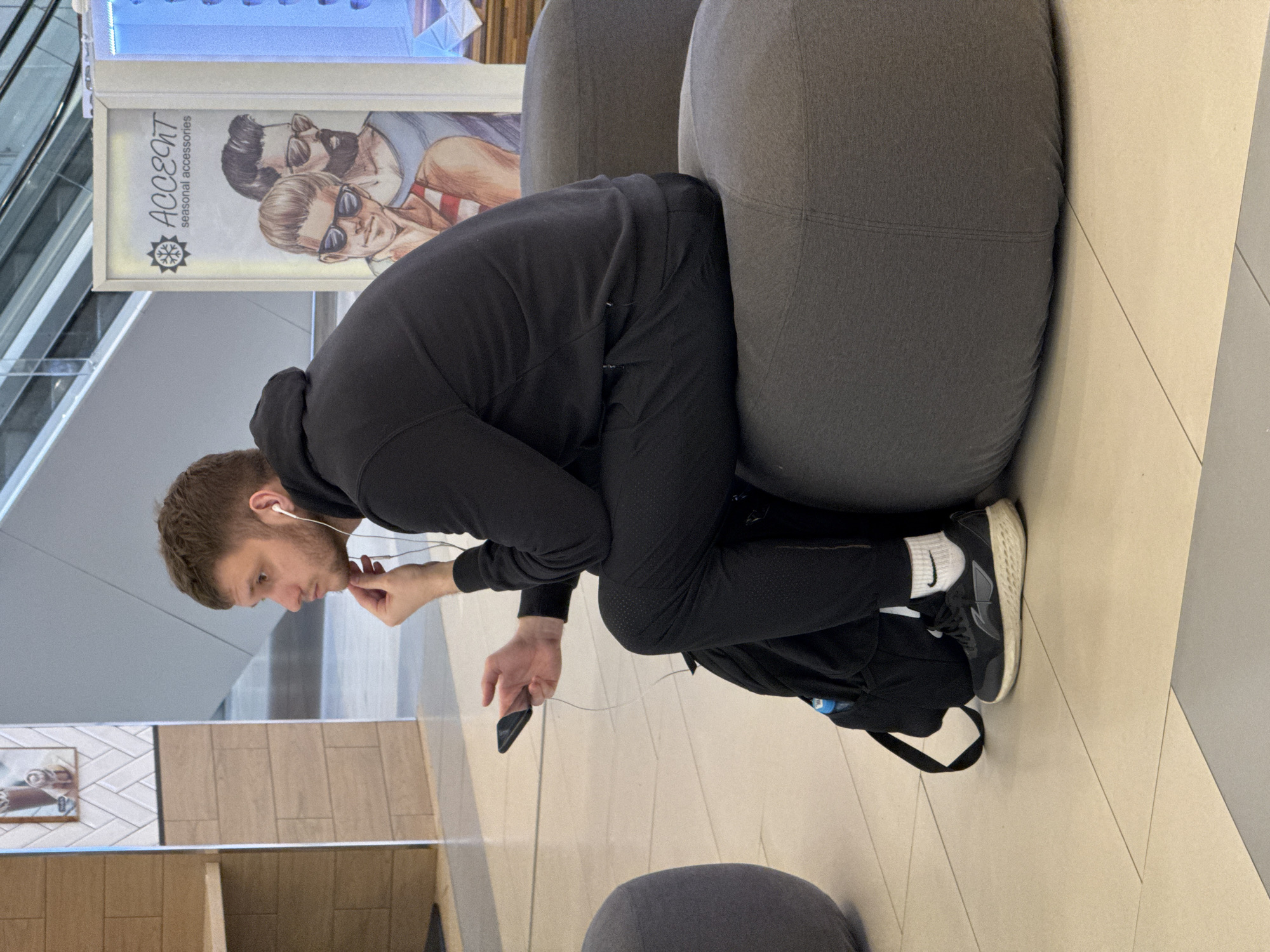 A man is squatting on a grey beanbag chair. He has headphones in and is holding a black cell phone in his left hand. He is wearing a black hooded sweatshirt, black pants, white socks, and black sneakers. There is a sign with the word "Accent" on a wall behind him, and a picture in a frame to the left. The floor is beige tile. The location is an interior space, potentially an airport or shopping mall in Saint Petersburg, Russia.