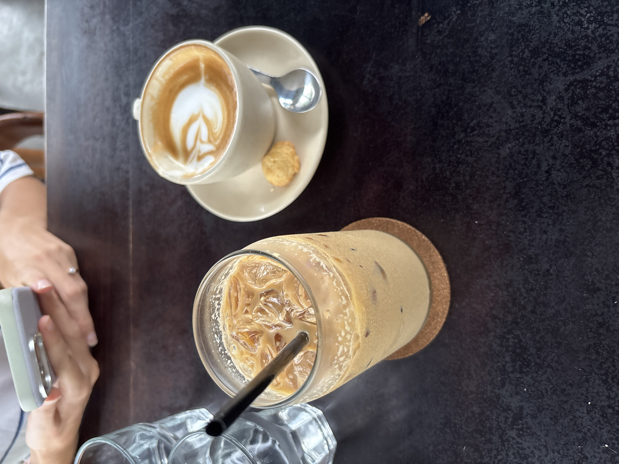 This overhead shot captures a dark, textured table with two coffee beverages and a person's hands. In the upper right quadrant, a light-colored ceramic cup holds a hot coffee featuring intricate latte art, resembling a heart or tulip pattern. It rests on a matching saucer, accompanied by a small silver spoon and a single, small golden cookie.
Below and slightly to the right, a tall glass of iced coffee stands, filled with ice cubes and a black straw. Condensation droplets are visible on the outside of the glass, which sits on a round cork coaster. Partially visible at the bottom left is an empty, clear glass with faceted sides.
To the left, a person's hands are visible, holding a light-colored rectangular object with a silver clip, possibly a phone or small electronic device. A silver ring adorns the ring finger of the left hand. The dark table surface appears mottled or speckled. The scene suggests a relaxed moment at a cafe, likely during the daytime. There is no visible text. The location context provided for this scene is Kuta Selatan, Indonesia.