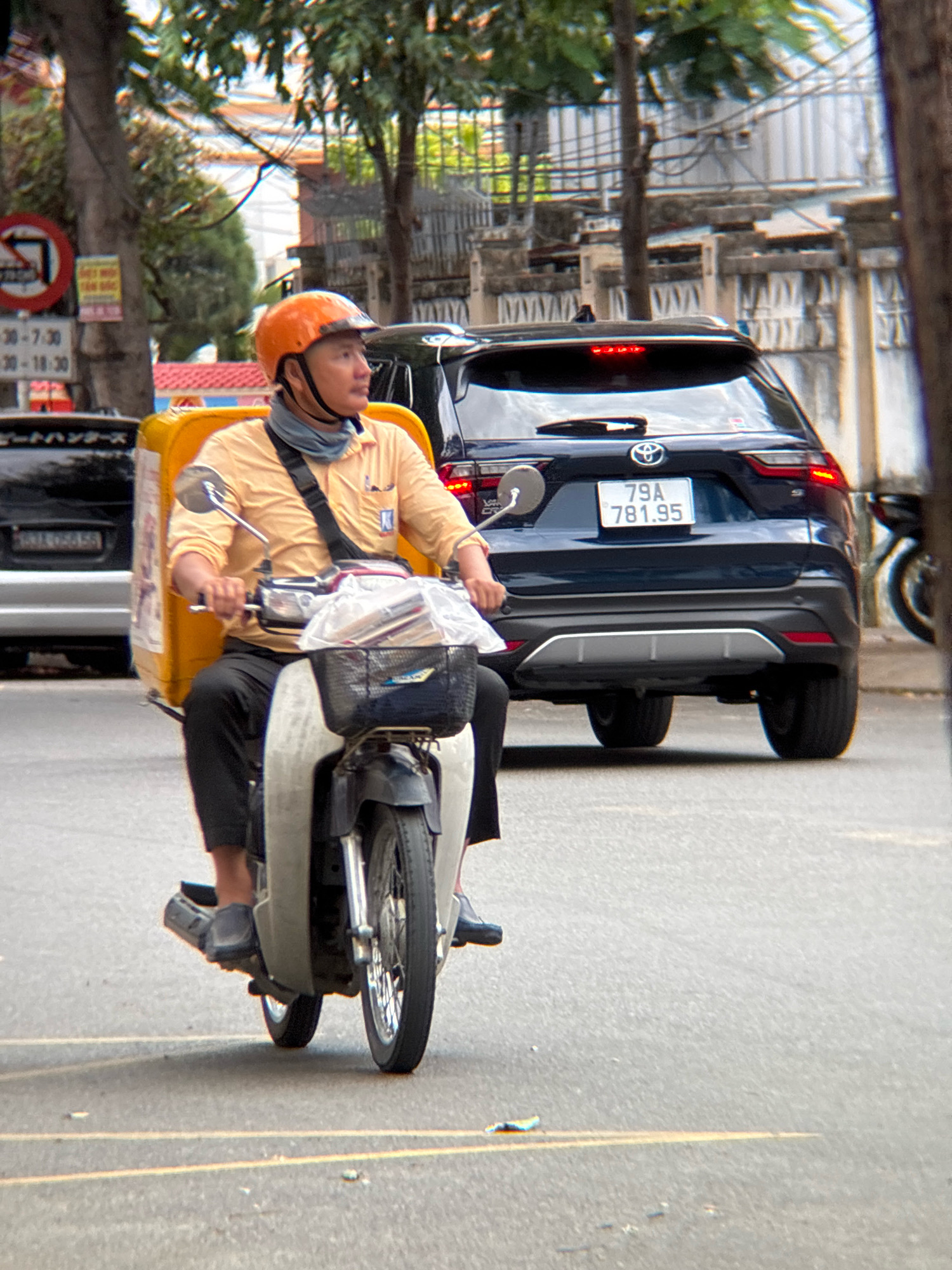 A person wearing an orange helmet and a light yellow shirt is riding a white scooter on a paved road in Nha Trang, Vietnam. The scooter has a large yellow storage box attached to the rear. The person is wearing black pants and black shoes. In the background, a dark blue SUV with a license plate reading "79A 781.95" is parked. There are trees and buildings visible, as well as street signs. The road has yellow lane markings. The lighting suggests it is daytime.