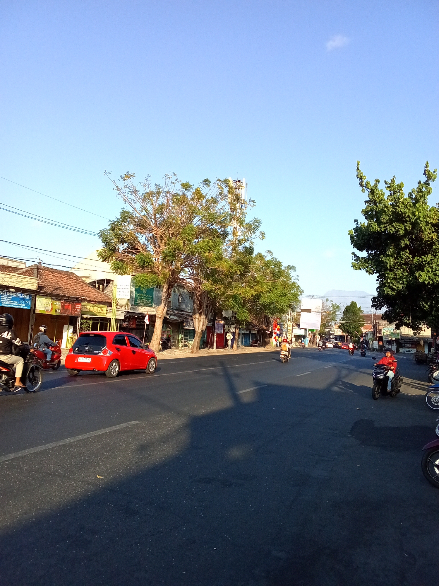 A street scene in Kediri City, Indonesia, features a bright, clear blue sky with a faint wisp of a cloud. The road is paved and appears to be a multi-lane thoroughfare. On the left side of the road, several low-rise buildings with tiled roofs line the street, some displaying signage. Trees with green foliage are prominent throughout the scene, with a large tree on the right casting a long shadow across the road.
Several vehicles are present. A red hatchback car is driving in the left lane. On the right side of the road, multiple motorcycles are visible, some with riders. One motorcycle rider in a red jacket is seen in the foreground on the right. Another motorcycle with a rider in a black jacket is in the left lane, moving away from the camera. Other vehicles, including more motorcycles, are further down the road, suggesting moderate traffic. The overall impression is a snapshot of daily life on a street in an Indonesian city during daylight hours.