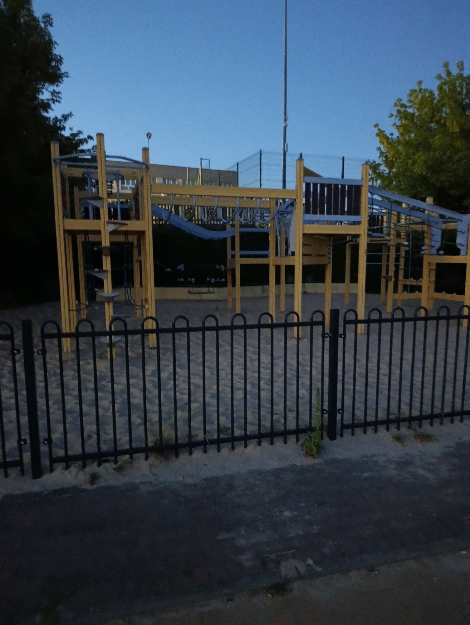 An outdoor view of a children's playground structure made of wood and metal is seen at dusk. The playground features multiple levels, slides, climbing nets, and bars. It is situated on a sand-covered area, enclosed by a black metal fence. A paved walkway is visible in the foreground. The sky is clear and blue, with a tall pole and some trees visible in the background.